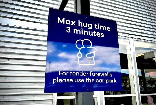 A sign at a new Zealand airport with hugging figures reading max hug time 3 minutes. For fonder farewells, please use the car park