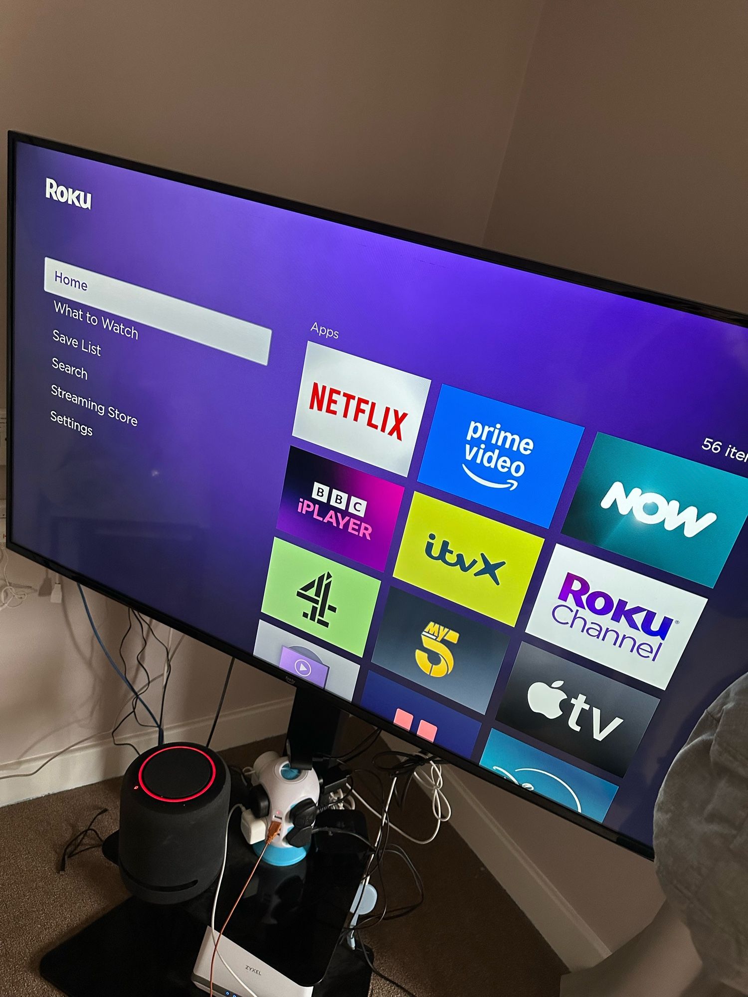 The image shows a television screen displaying the Roku home interface. The background is purple, and on the left side, there is a menu with options: Home, What to Watch, Save List, Search, Streaming Store, and Settings. On the right side, there is a grid of app icons. The visible apps include Netflix, Prime Video, Now, BBC iPlayer, ITVX, Channel 4, My5, Roku Channel, and Apple TV. Below the TV, there is a black cylindrical speaker with a red ring on top, some cables, and a small white and blue device that looks like a toy or a gadget. The TV is mounted on a stand, and there are various cables connected to it.