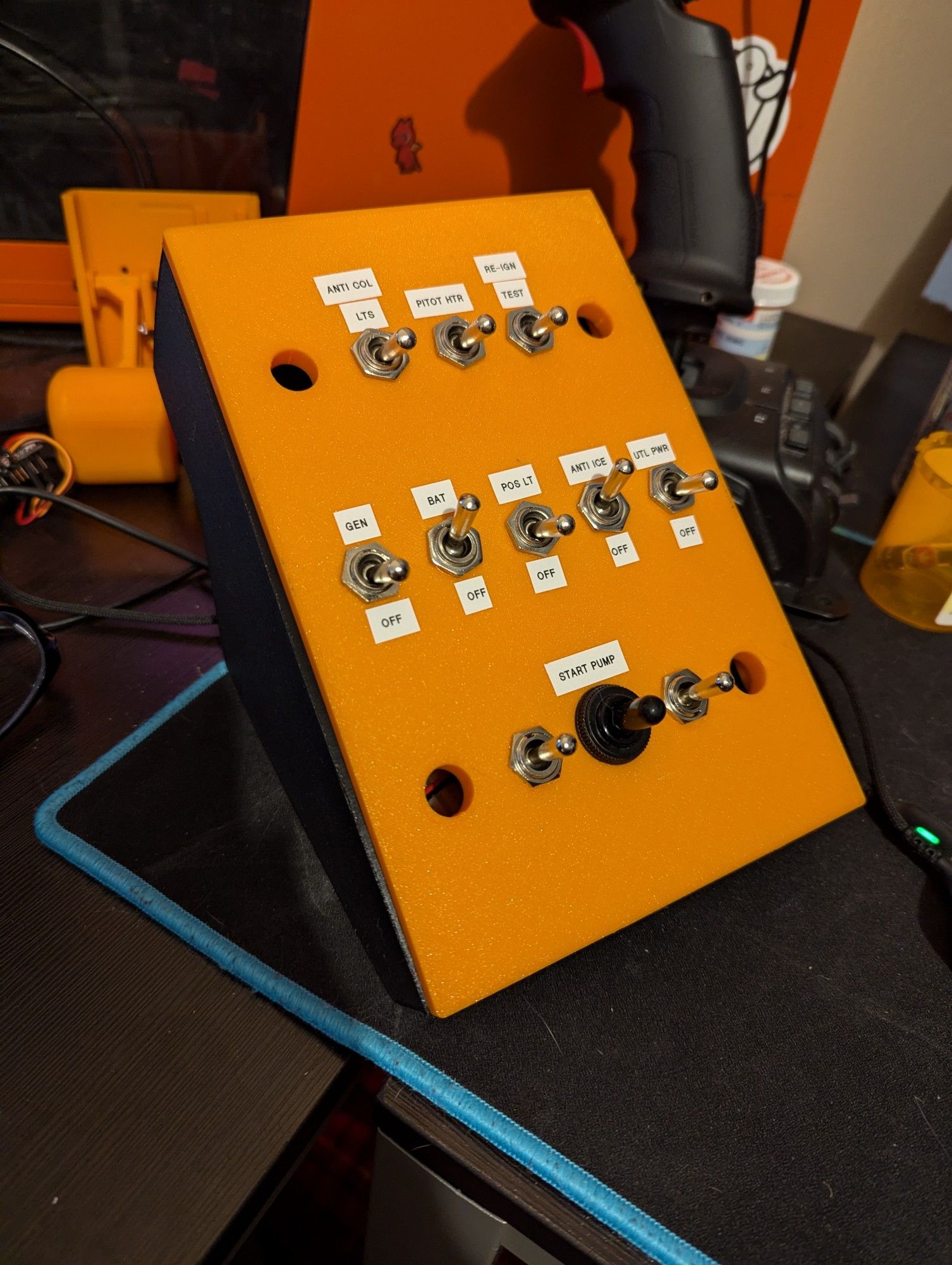 An orange 3d printed panel mounted on a black 3d printed housing.
The panel is inclined at around 45 degrees and has 15 holes in it for switches. Most of the switches have been installed and are labelled for various functions in a helicopter cockpit.
The panel is intended for use in flight simulators.