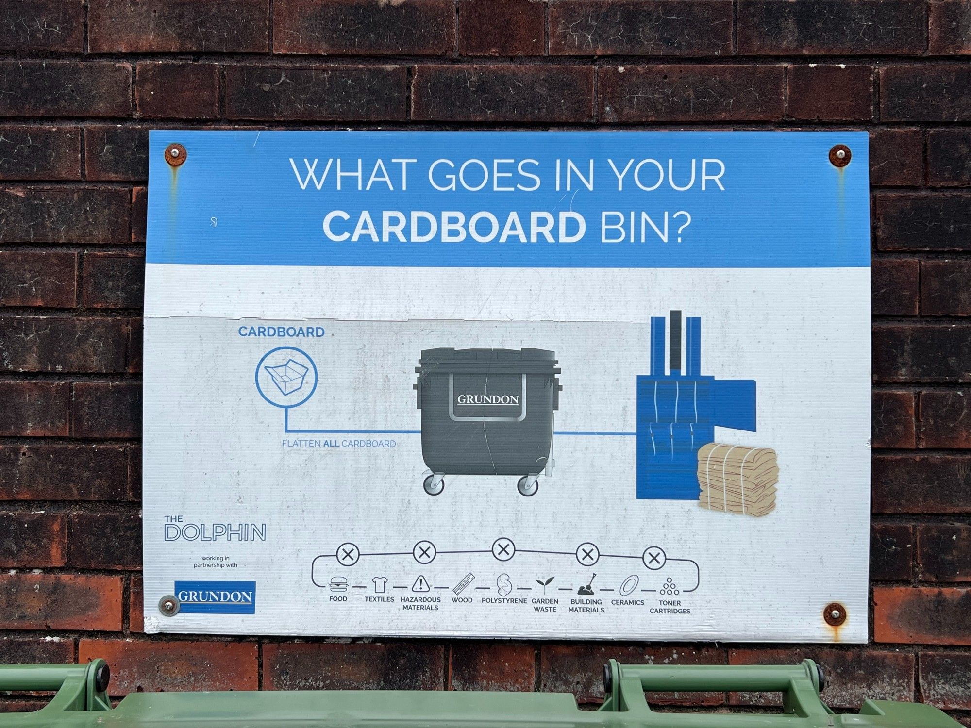 A sign above a bin for cardboard stating that cardboard should go in the bin.