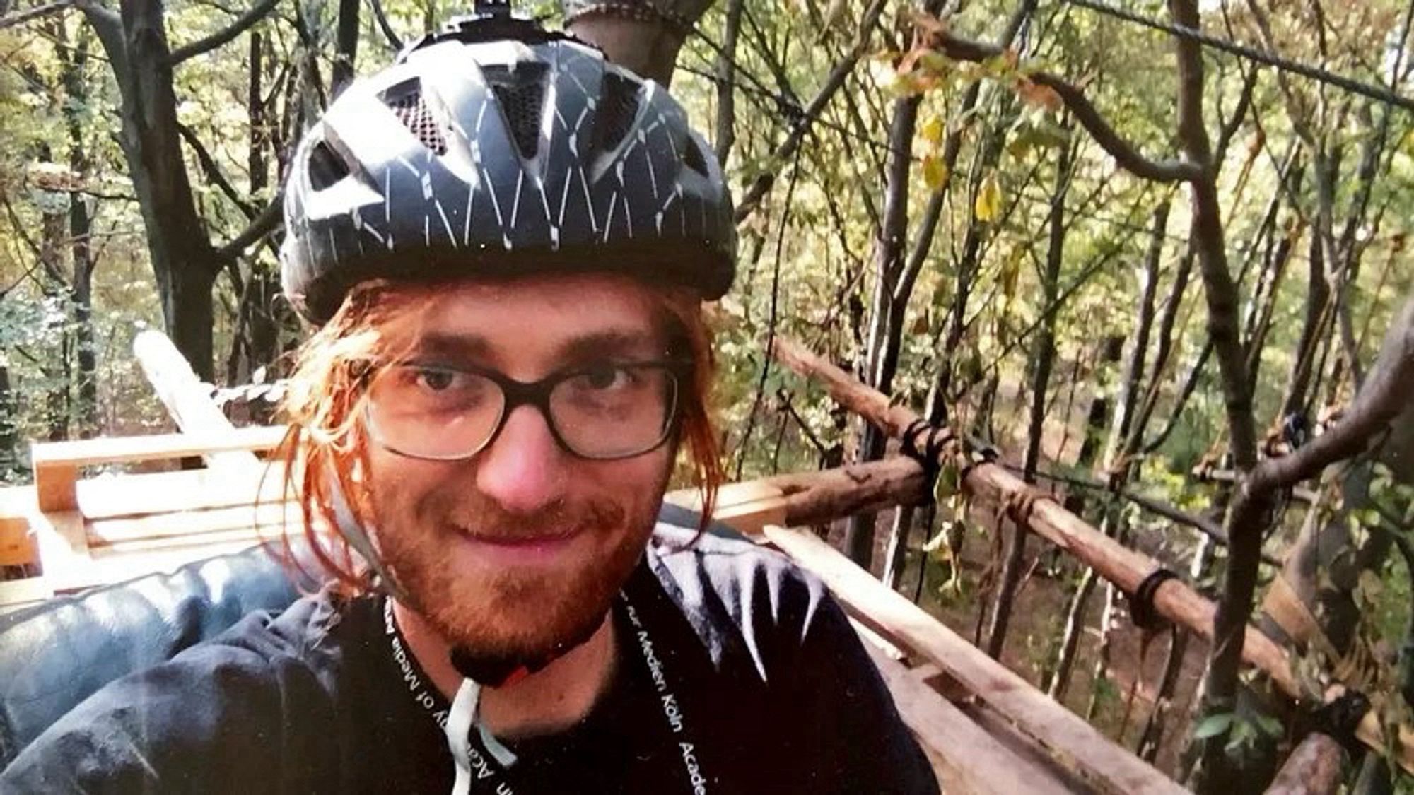 Selfie von Steffen Meyn: Mensch mit roten langen Haaren und rotem Bart. Trägt Brille mit großen Gläsern und schwarzem Rand. Trägt einen schwarzen Fahrradhelm und ein schwarzes T-Shirt. Im Hintergrund sieht eins eine in den Bäumen gebaute Plattform aus Holz, auf der der Mensch steht. Der Mensch lächelt in die Kamera.