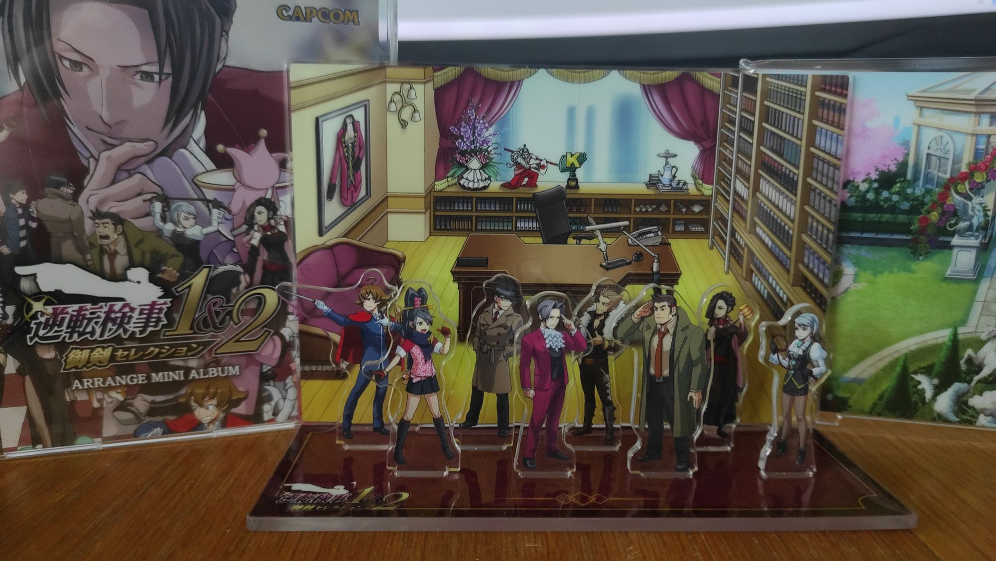 Diorama fourni avec l'édition limitée de Miles Edgeworth Investigations. Huit personnages du jeu sont posés sur un support acrylique avec l'un des deux fonds fournis (ici le bureau), arrangés de façon à donner l'impression de discuter. Un individu suspect n'ayant pas l'air de suivre la discussion regarde ailleurs. Peut-être le mur, ou peut-être vers le CD de l'ost réarrangée juste à côté.