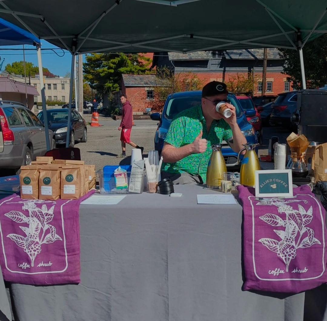 Co-owner Justin, a white man in a green shirt drinks from a white paper coffee cup. He is standing behind a table with a grey tablecloth and two purple coffee sacks. Bags of coffee sit to the left and carafes of coffee sit to the right.