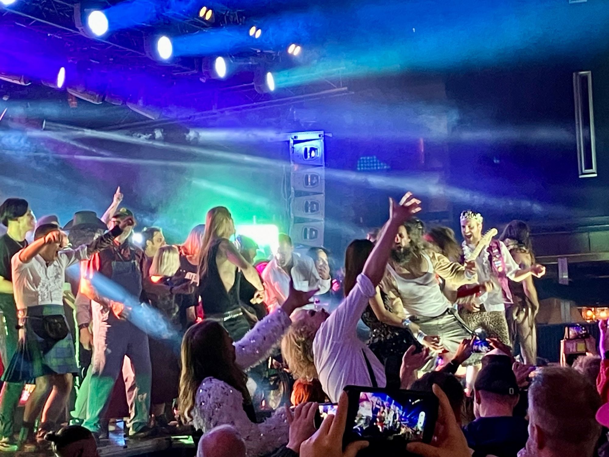 Several people in various states of hard rock undress, play air guitar on a stage while a the audience presses in close with hands and phones in the air. The light is bluish green with beams of stage lights illuminating the crowd.