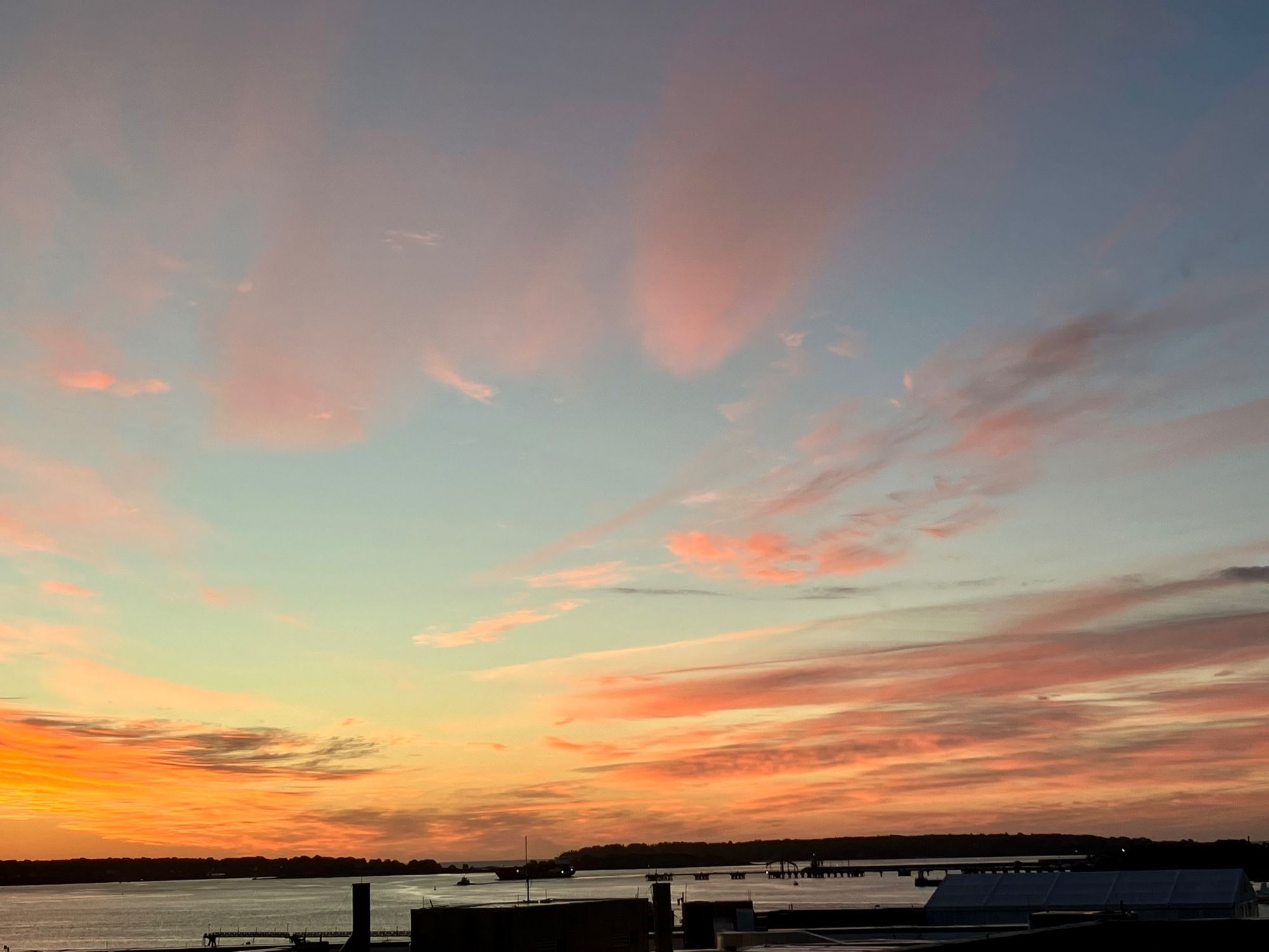 Image of the sunrise outside my hotel window in Portland, Maine. There are several pink clouds, some streaky and some fluffy. The sky is orange and blue and there is a small container ship in the blue-grey water.