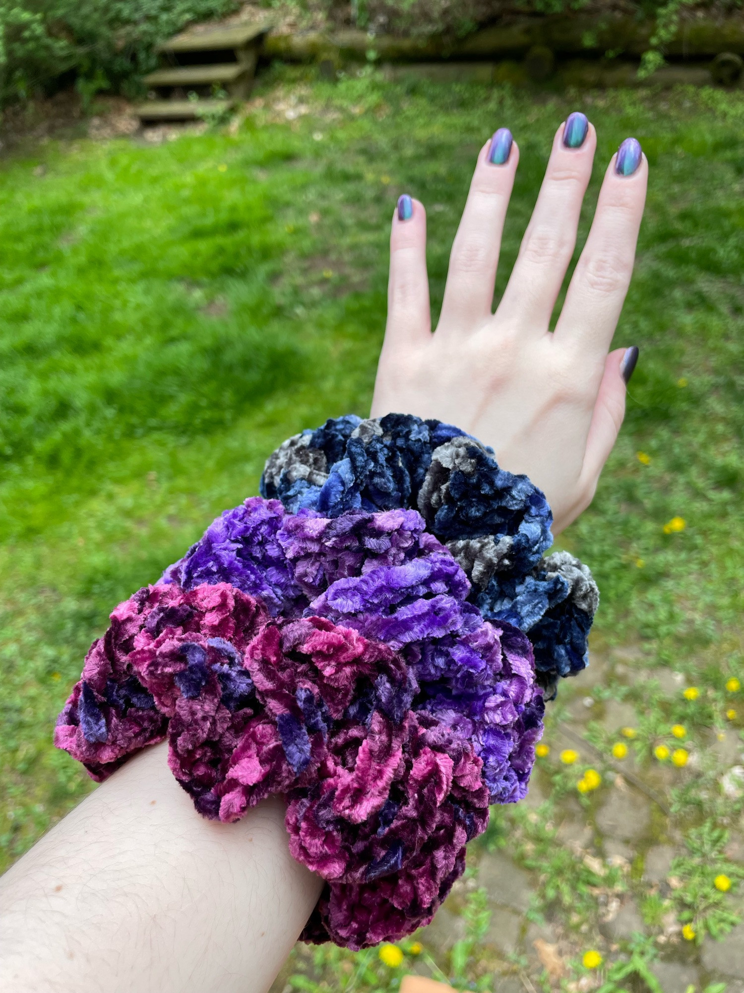 Jenna’s arm showing off crushed velvet navy, crushed velvet purple, and crushed velvet burgundy scrunchies outside in her backyard, there is mostly grass in the background. Jenna’s arm is pale, turned palm facing down, back of hand up with fingers stretched out, and showing off a purple green multichrome nail polish