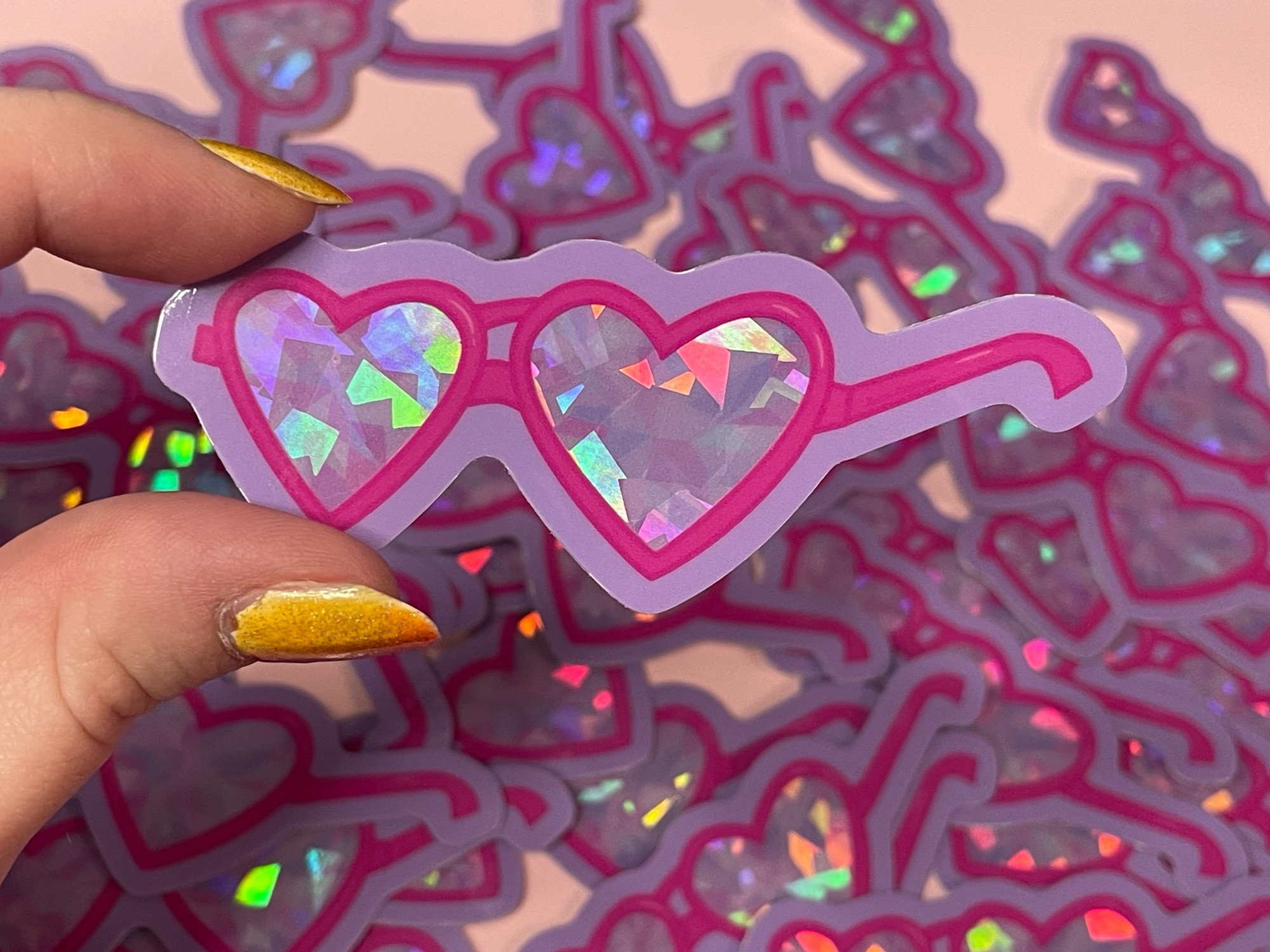 Jenna holding her sticker above a pile of stickers of the same design on a light pink desk in the background. Sticker design its heart shaped glasses with pink frames, lenses with fractal shapes in pinks and purples with a prism holo effect over them, and a light muted purple border around the whole design.