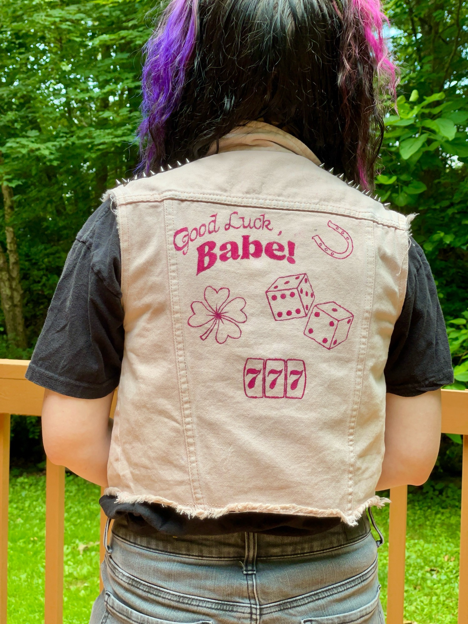 Jenna wearing a vest she painted with her Good Luck Babe inspired design. The vest is lights pink denim, the pint is fuschia. It says Good Luck and Babe in 2 retro fonts on the upper left corner or the center of the back panel of the vest. To the right is a horseshoe, black is a four leaf clover and a pair of dice, below that is a luck 777 slot. Under the vest she wears a black t shirt, and wears gray pants. Her hair is black in half up pigtails split dyed purple and pink. She is outside, behind her visible is a light brown fence for her deck, green grass, trees and other greenery. 