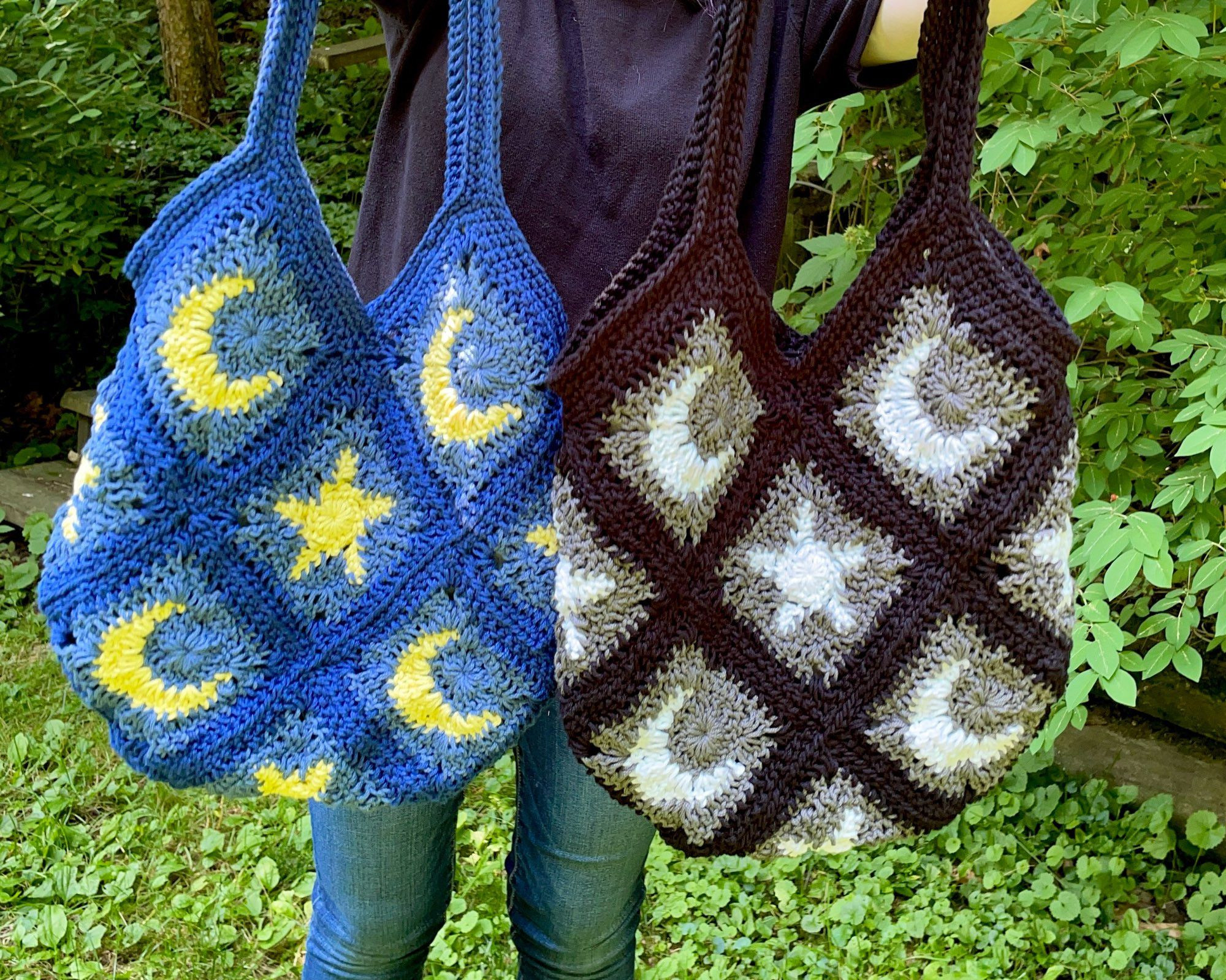 The 2 variations of the night sky moon & star bag, blue & yellow left, black, gray & white right. The squares are laid out diagonal, moons top & bottom, stars middle & base. They're being held by the artist outside surrounded by greenery