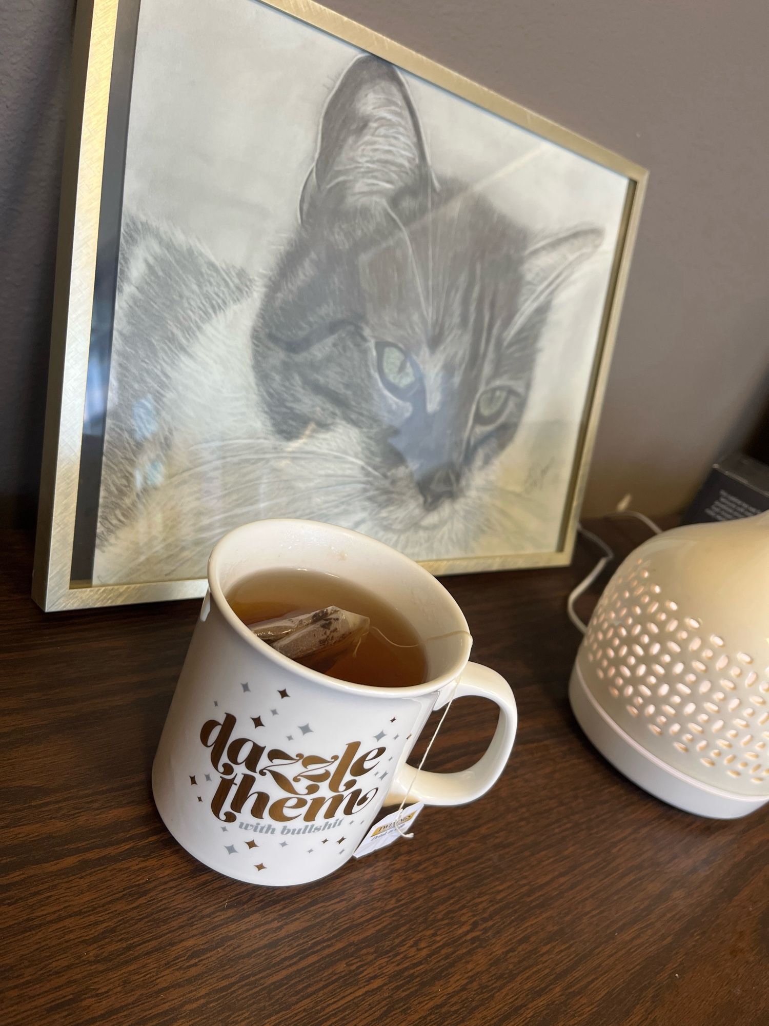 A teacup that says “Dazzle them with bullshit” on a desk in front of a framed sketch of a cat. Some might recognize the cat as Persephone.