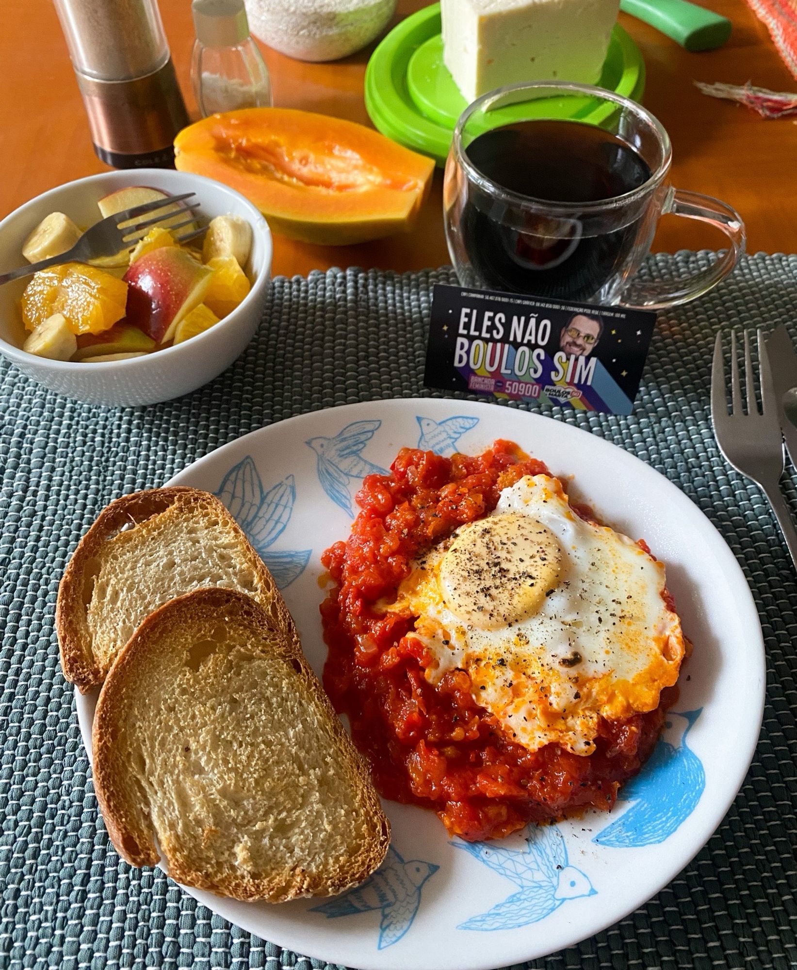 Mesa de café da manhã: prato com 2 fatias de pão torrado e shakshuka (molho de tomate com um ovo por cima), xícara de café, pote com frutas picadas, mamão papaia cortado ao meio, queijo, saleiro, pimenteiro e pote com aveia. Apoiado na xícara, tem um cartãozinho com os dizeres “ELES NÃO, BOULOS SIM”