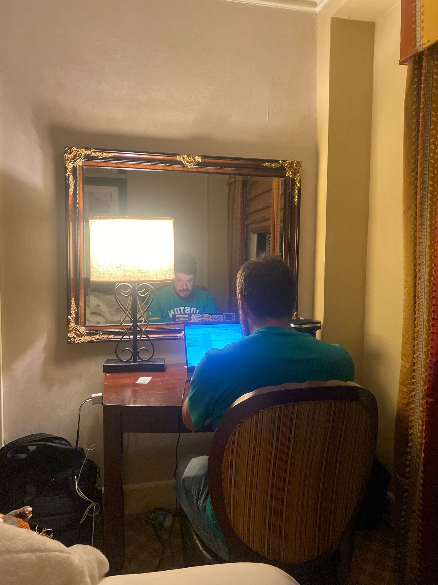 From behind, a man sitting at an ornate desk typing on a laptop. His face (bearded, Caucasian) reflected in the mirror above the desk. There is a lamp on the desk as well.