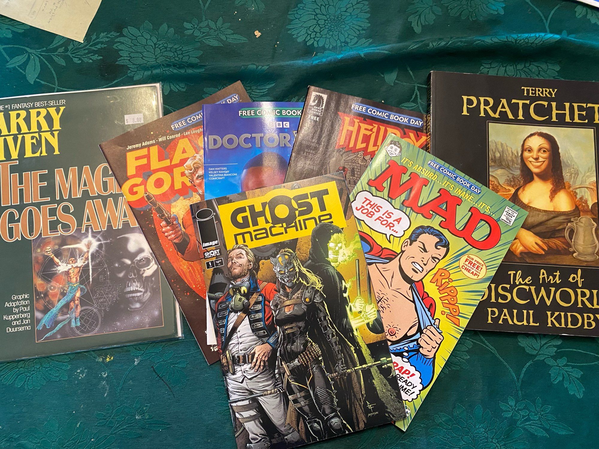 A bunch of comics on a table. Ghost int he machine, a mad magazine Superman parody, bellboy, Doctor who, “the Magic Goes Away” by Larry Niven, and The Art of the Discworld