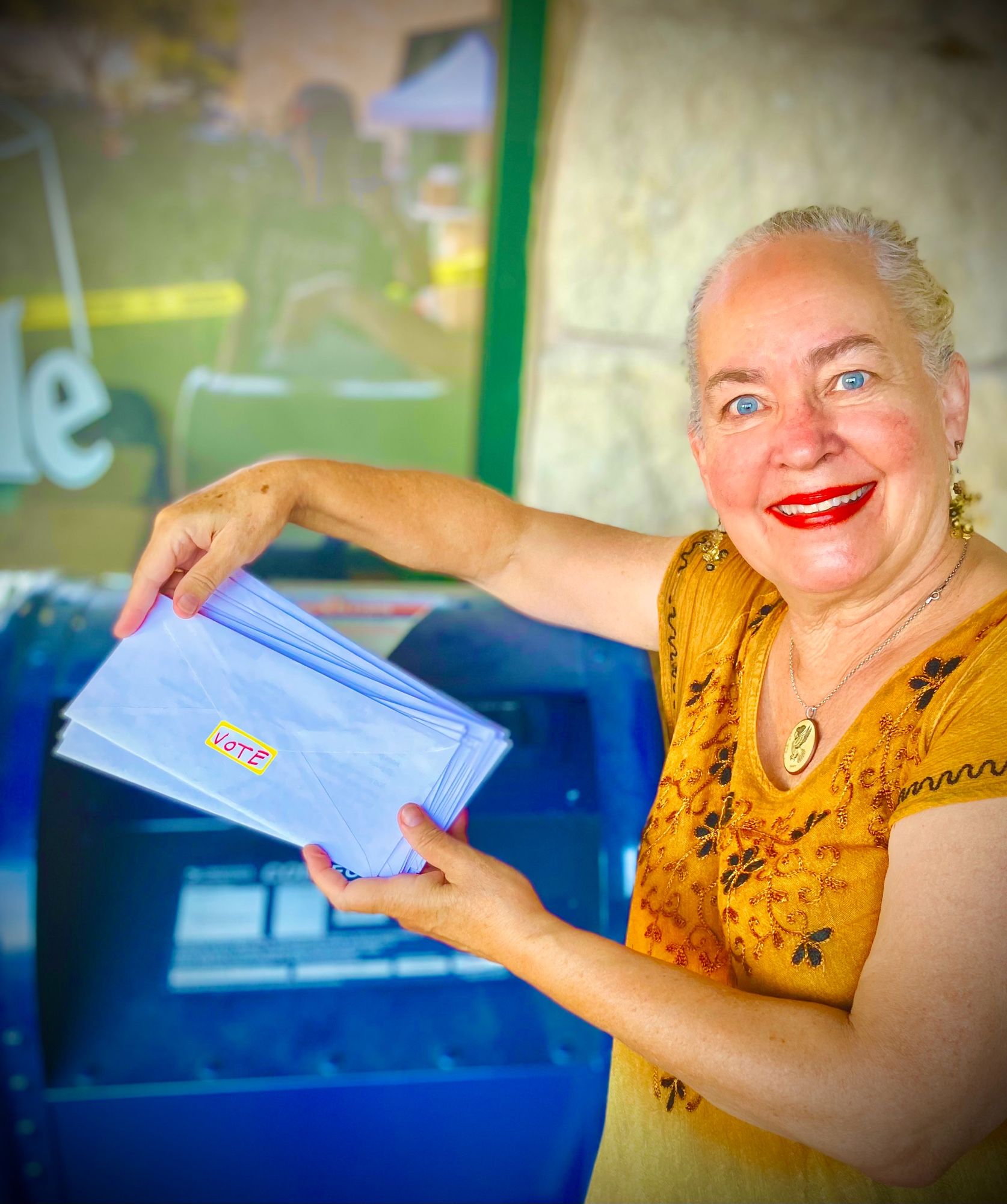 A photo of me about to put a batch of Get Out The Vote letters into an old-fashioned blue mailbox. The letters have a VOTE sticker on the back of them.
I’m an old, white-haired, blue-eyed,
white woman. I’m wearing a mustard colored embroidered sundress and bold red lipstick.