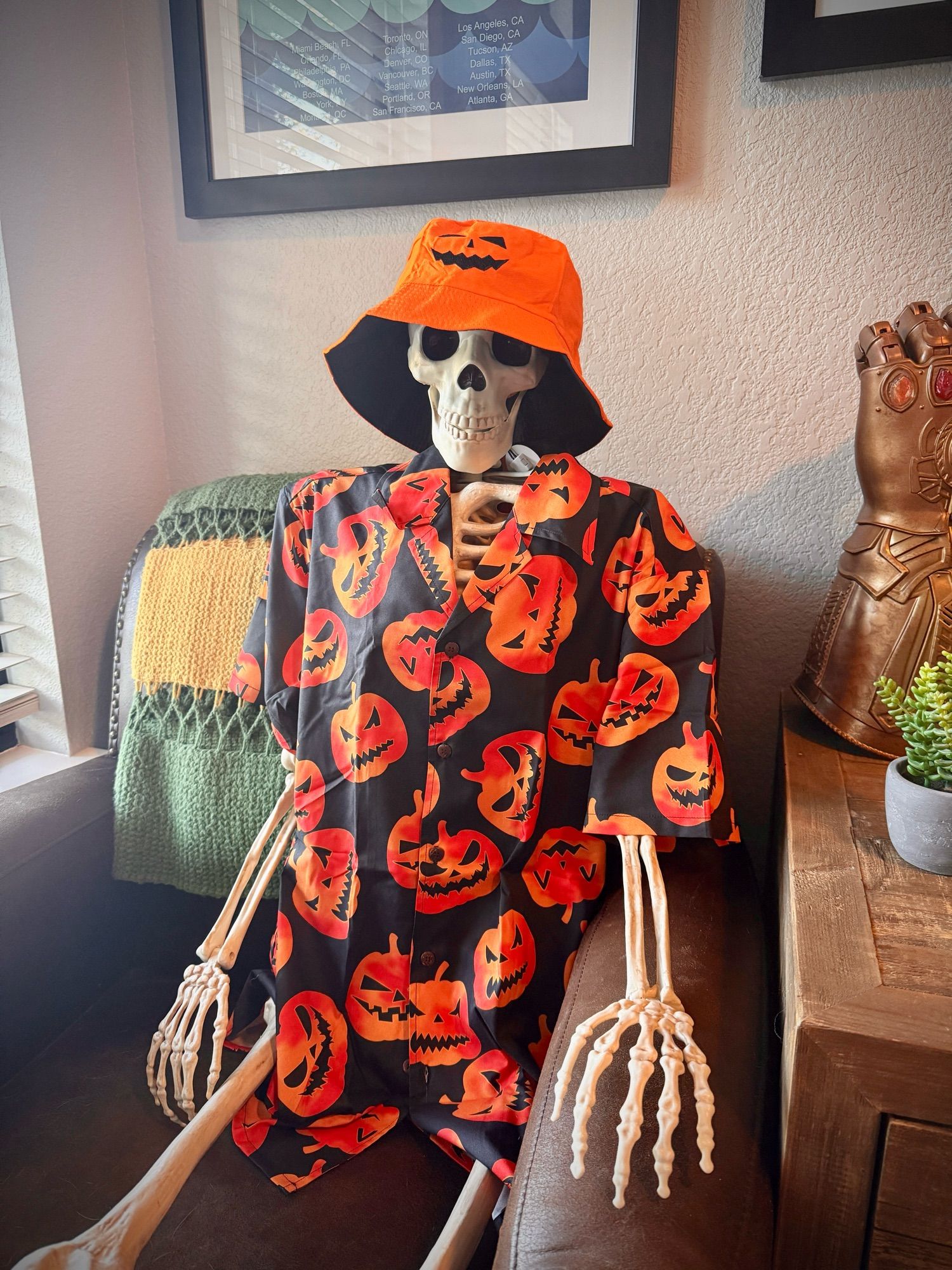 A photo of a life sized skeleton wearing a black button up shirt with orange Jack-o-lanterns all over it. He is also eating an orange bucket hat with a Jack-o-lantern on it.