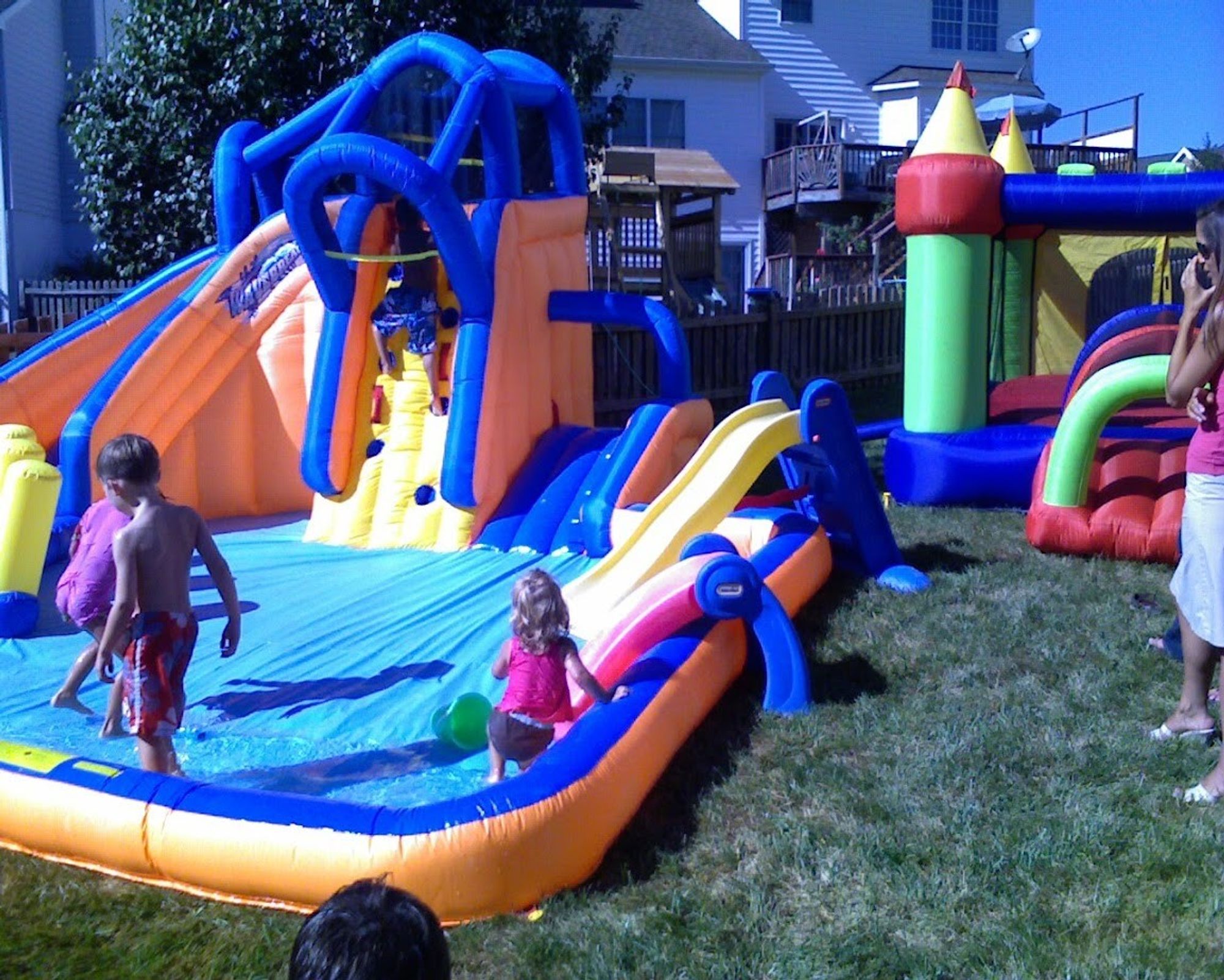 An orange & blue inflatable waterslide next to a primary colors moonbounce in a back yard with yound kids on them