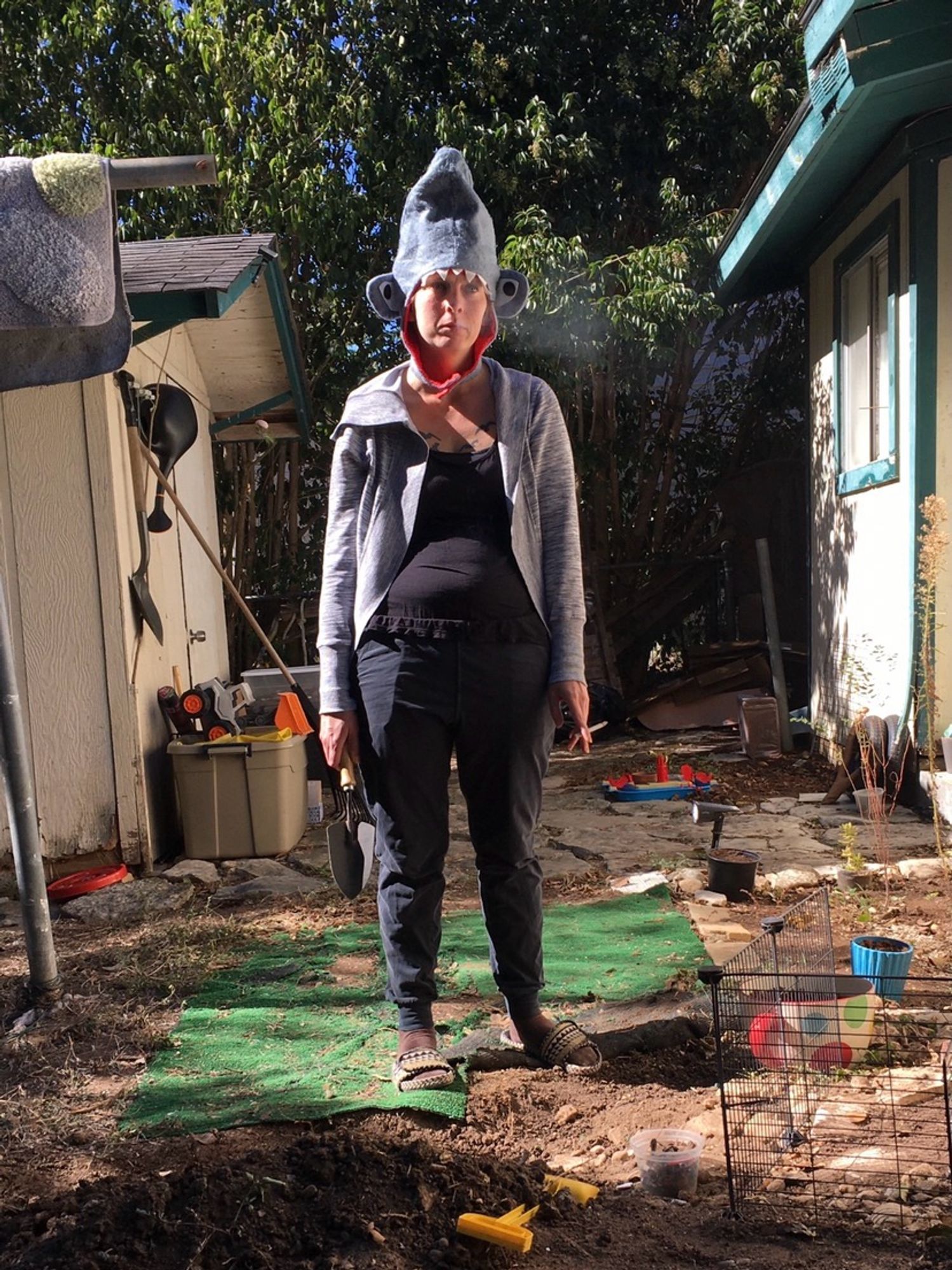 Image of a tired me holding a spade in a backyard that needs a lot of love. I am wearing my shark hat.