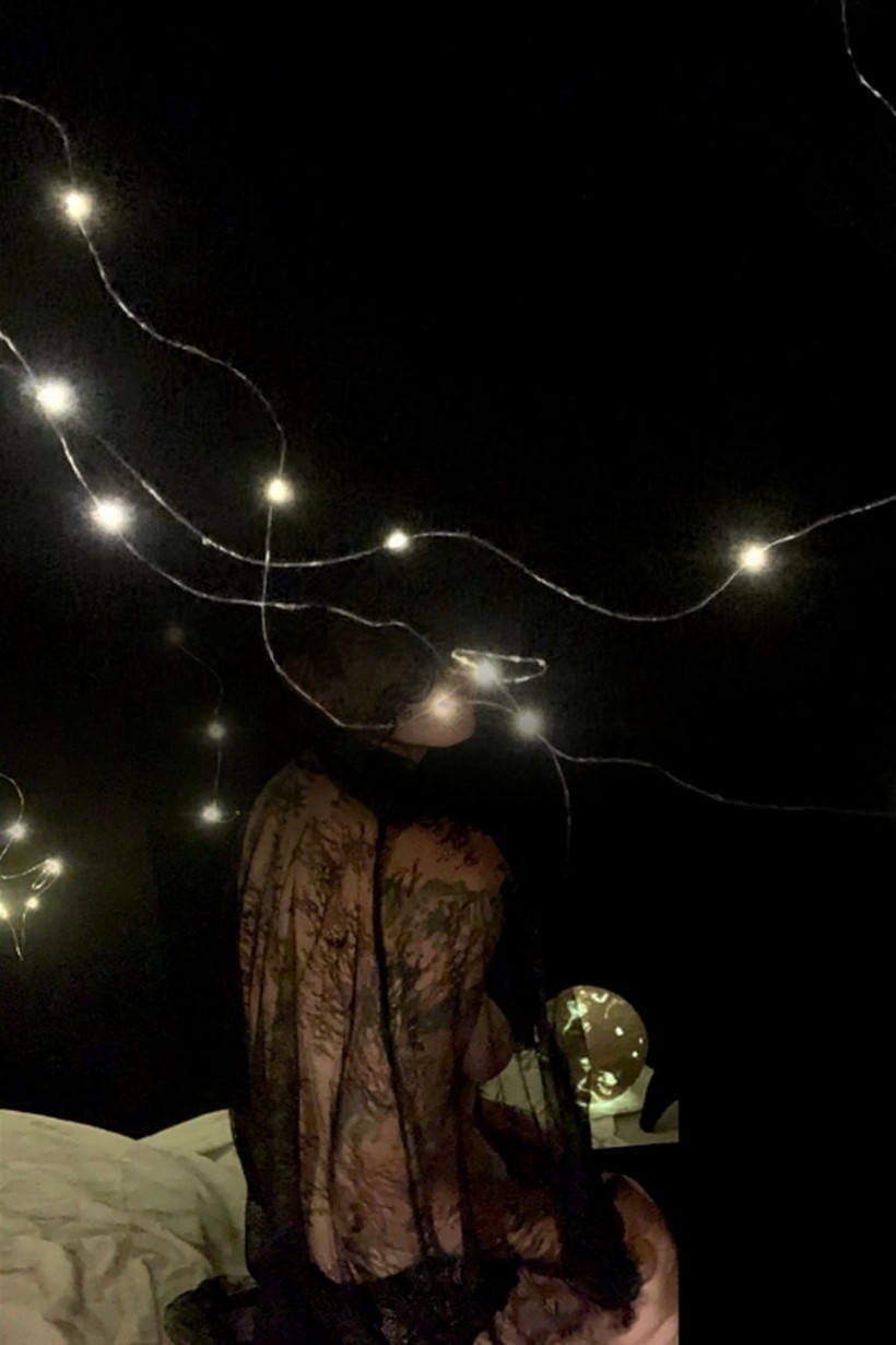 Photo of the artist sitting on their knees in a black lace cloak. The paper mache moon is on the ground behind her. The background is darkness and fairy lights behind the white fabric they are kneeling on. Photo is blended at the top with a photograph of fairy lights which come across the top and the artist’s face, making it look like they are disappearing. 