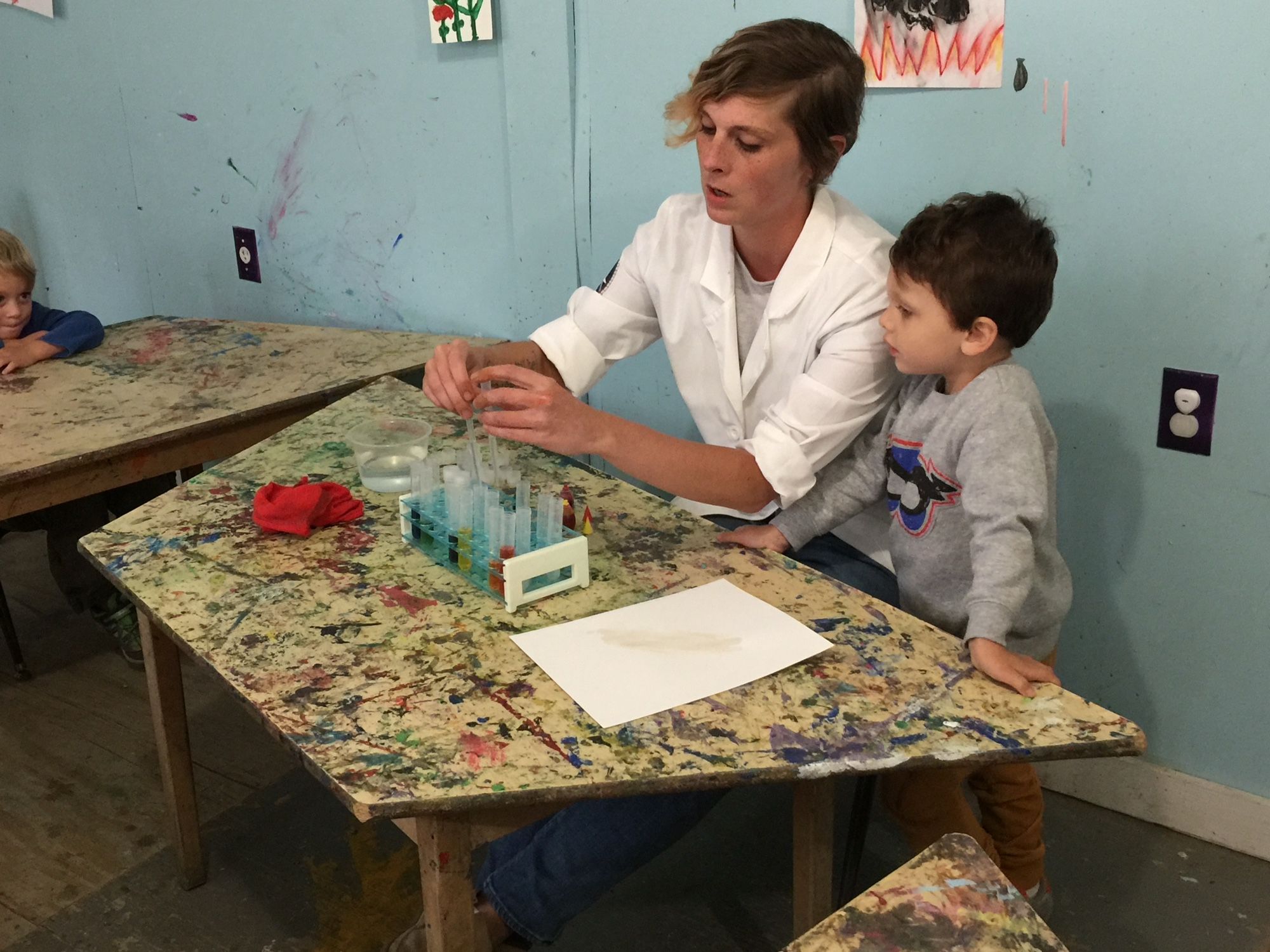 Photo of me teaching color mixing to young children. One child is pictured. I am using food coloring and water in a science looking beaker set up and wearing my labcoat.