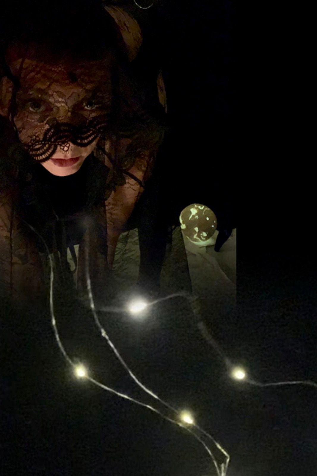 Artist leaning over and facing the camera with arms hanging down. The background is blackness except a paper mache moon that is lightly illuminated and on the ground behind them.
They are draped in black lace except for their mouth and chin. Photo is blended with a photograph of fairy lights which reach out from her chest and cloak and come forward toward the viewer at the bottom of the photo.