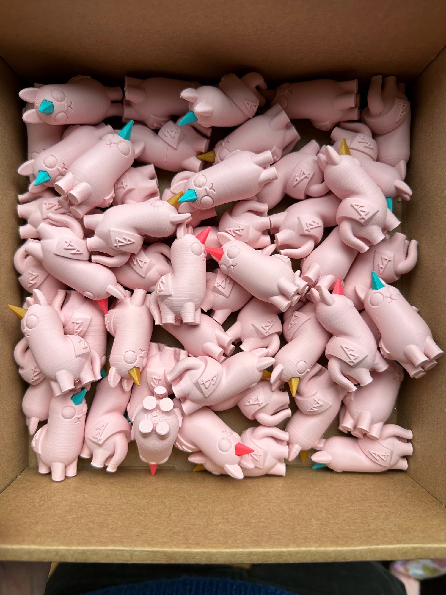 A box full of small baby pink plastic creatures with single horns in gold, hot pink or turquoise, large eyes, long ears and noses and mouths like a cat or rodent.