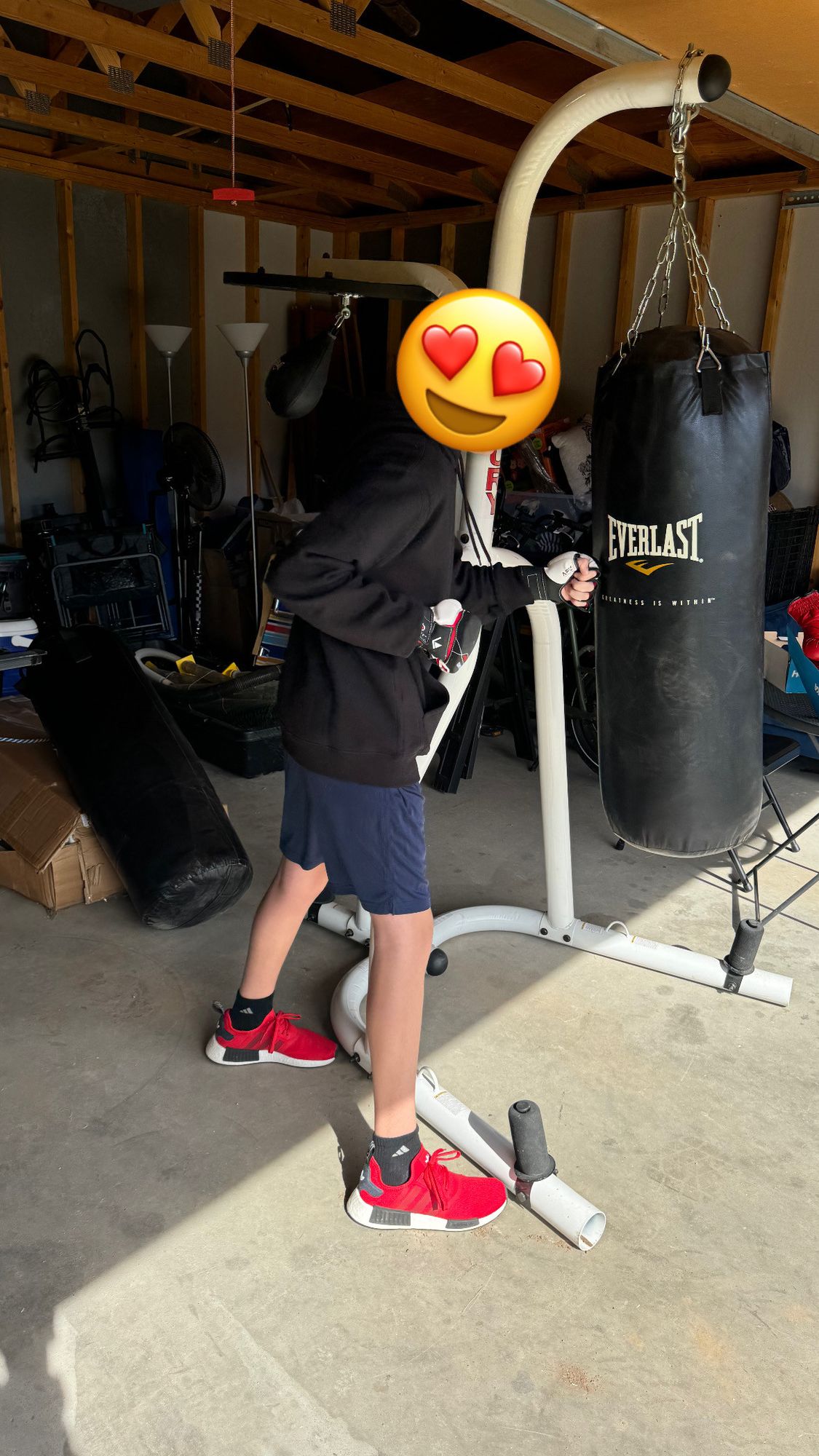 Photo of a teenager in a black hoody, blue shorts, and red shoes with their face covered punching a punching bag in a garage