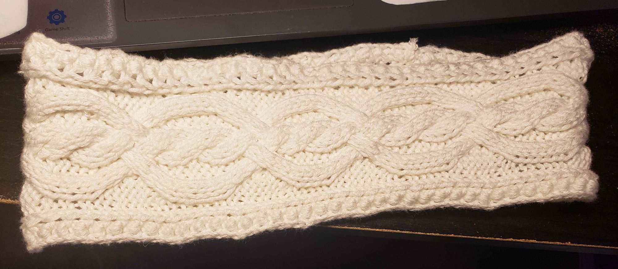 White cable headband laid flat on a desk. It has a twisting central cable pattern and a knotted border.