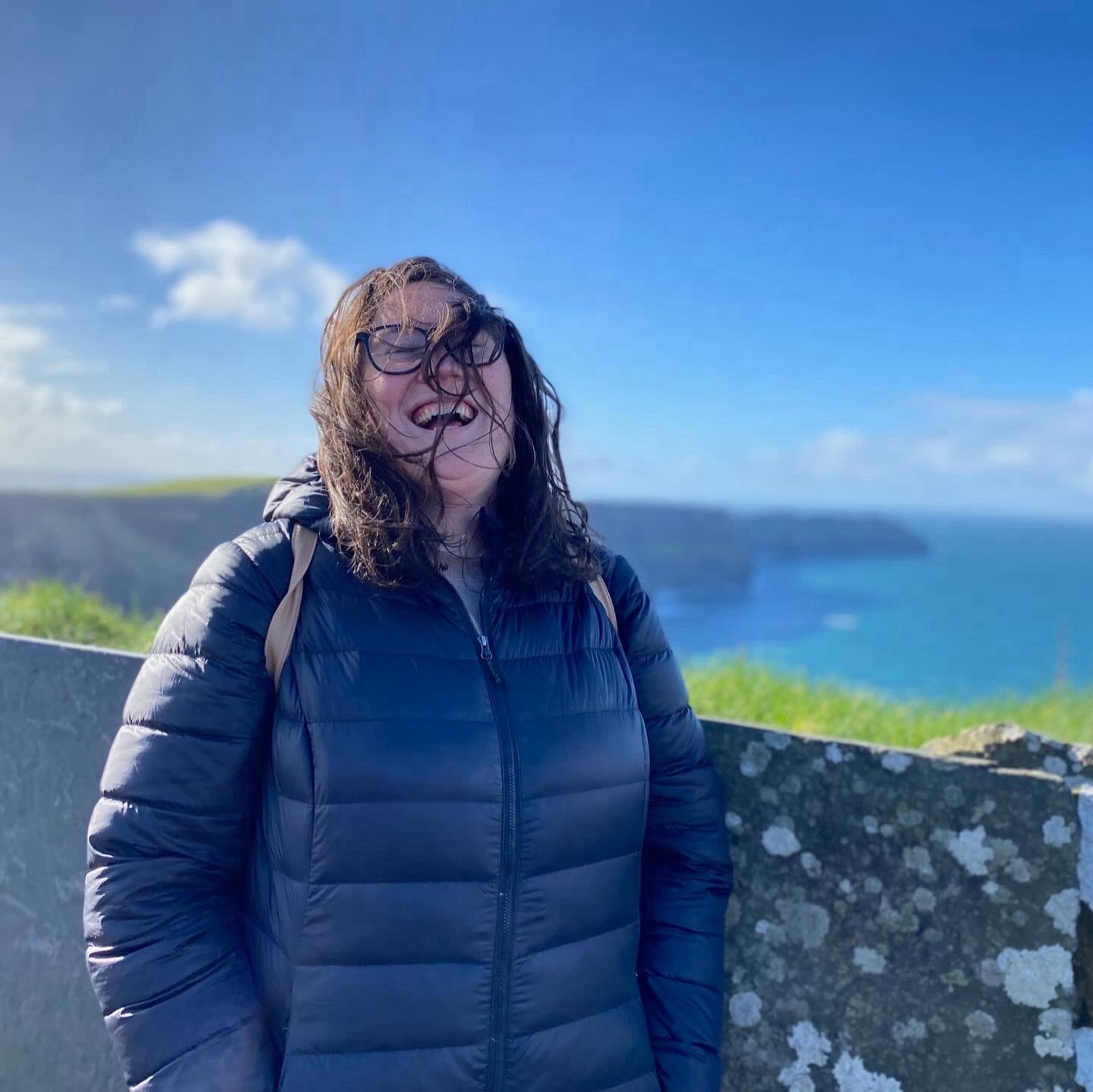 A woman in a black coat laughs as her hair blows in her face in front of the Cliffs of Moher