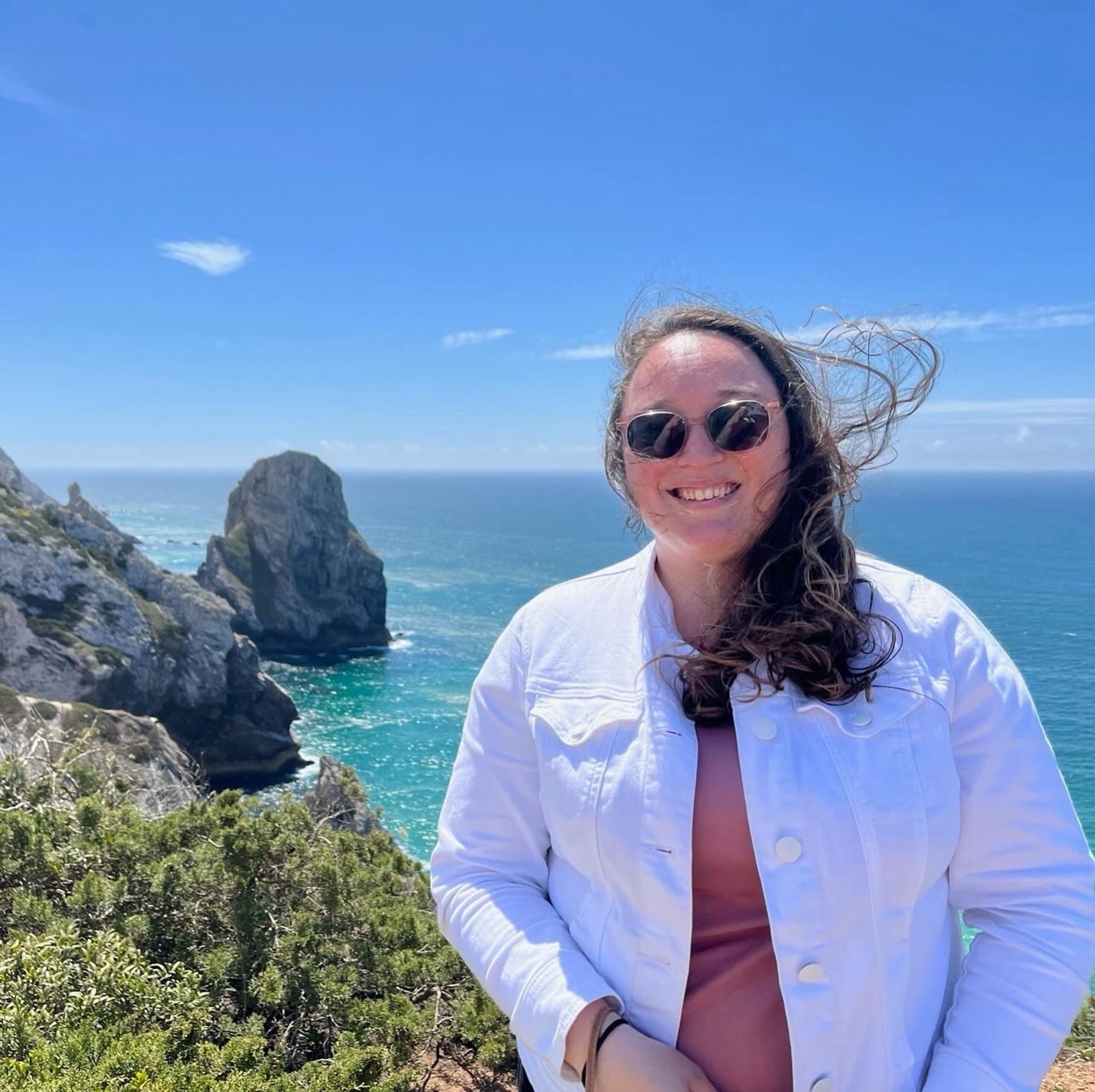 A woman in a white shirt has her hair blown with the ocean in the background