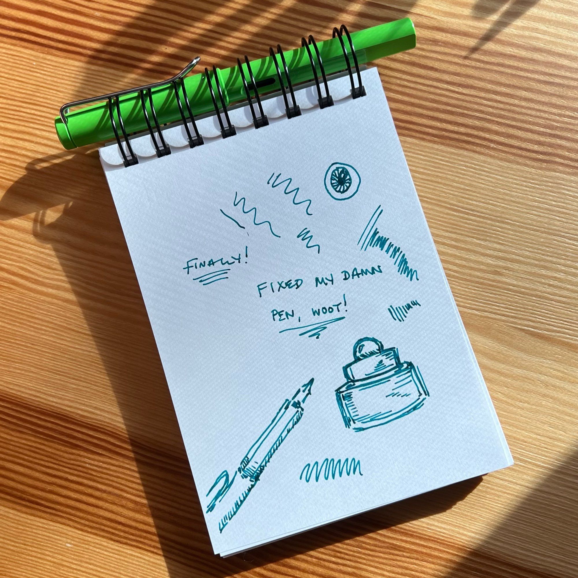 Small sketchbook page with teal green ink squiggles, doodles of a fountain pen and ink bottles, and writings of “Finally!” & “Fixed my damn pen, woot!”