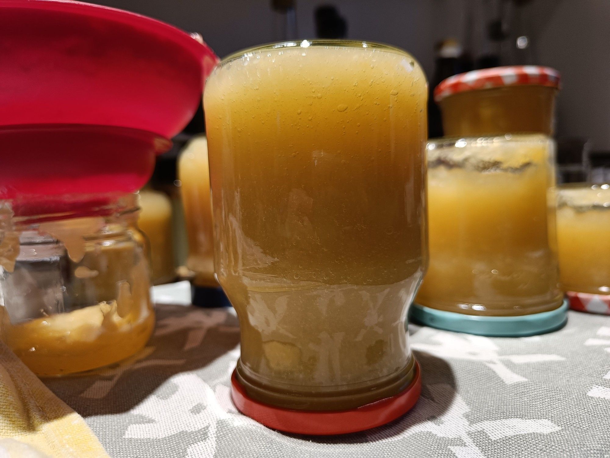 Auf dem Kopf stehendes Glas mit gelber Marmelade, weitere Gläser und ein roter Trichter unscharf an den Seiten 