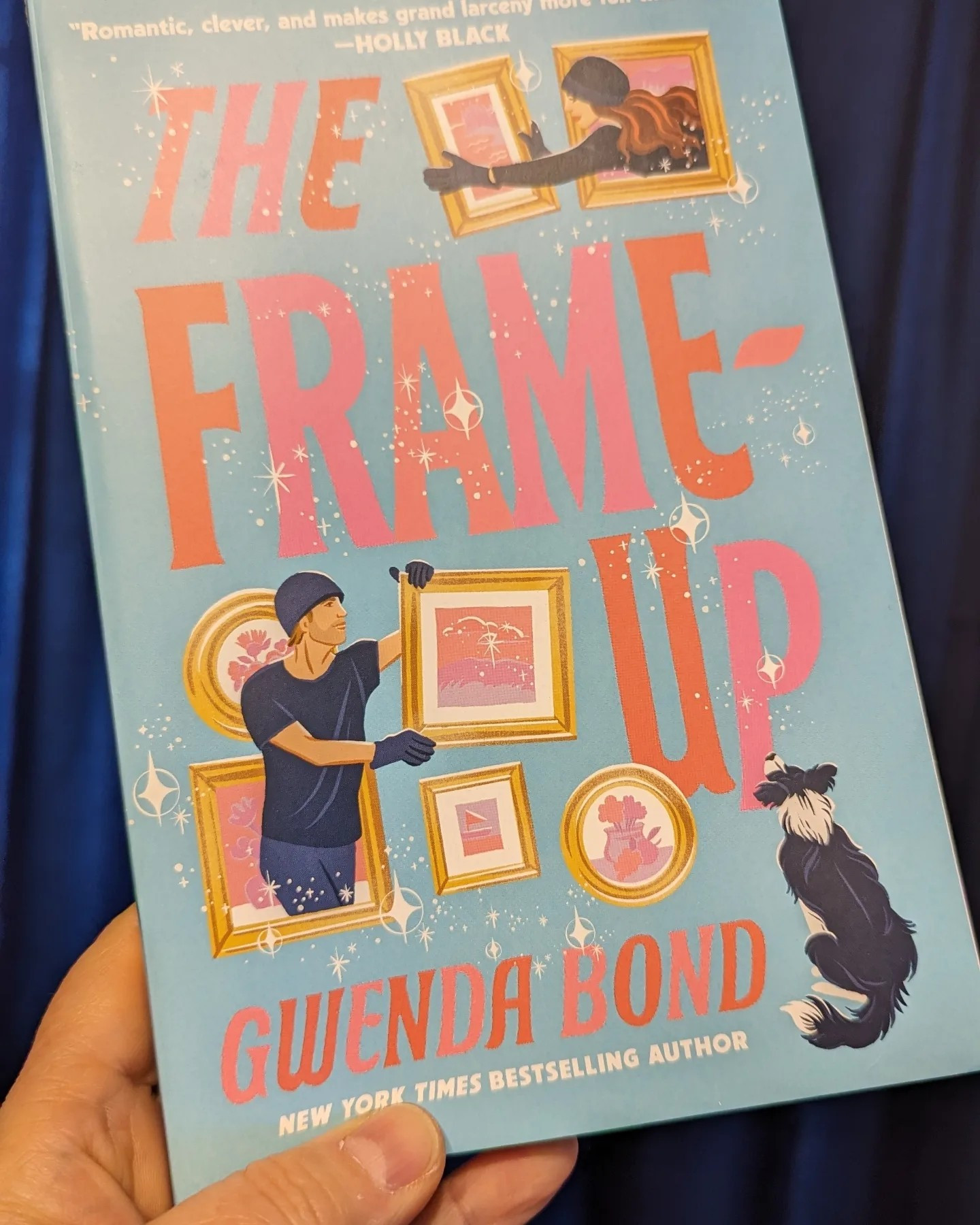 Cover of a book entitled The Frame-Up by Gwenda Bond. Blue background, illustration of a man and woman dressed in black as burglars grabbing picture frames off the wall while a black and white Border Collie sits watching.