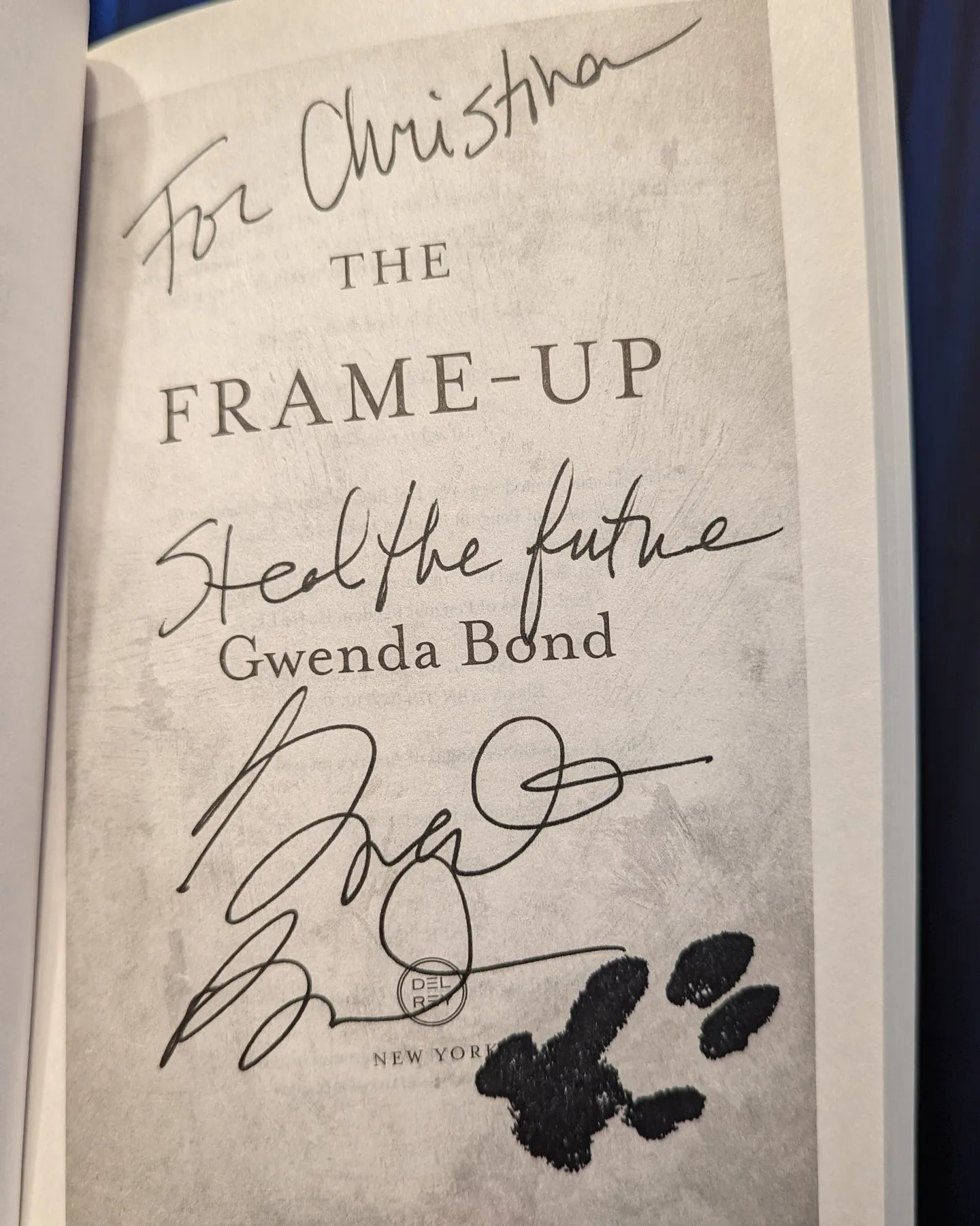 Title page of The Frame-Up by Gwenda Bond shows it autographed "to Christina, steal the future". Below the author's signature is a black ink stamp of a dog's paw print.