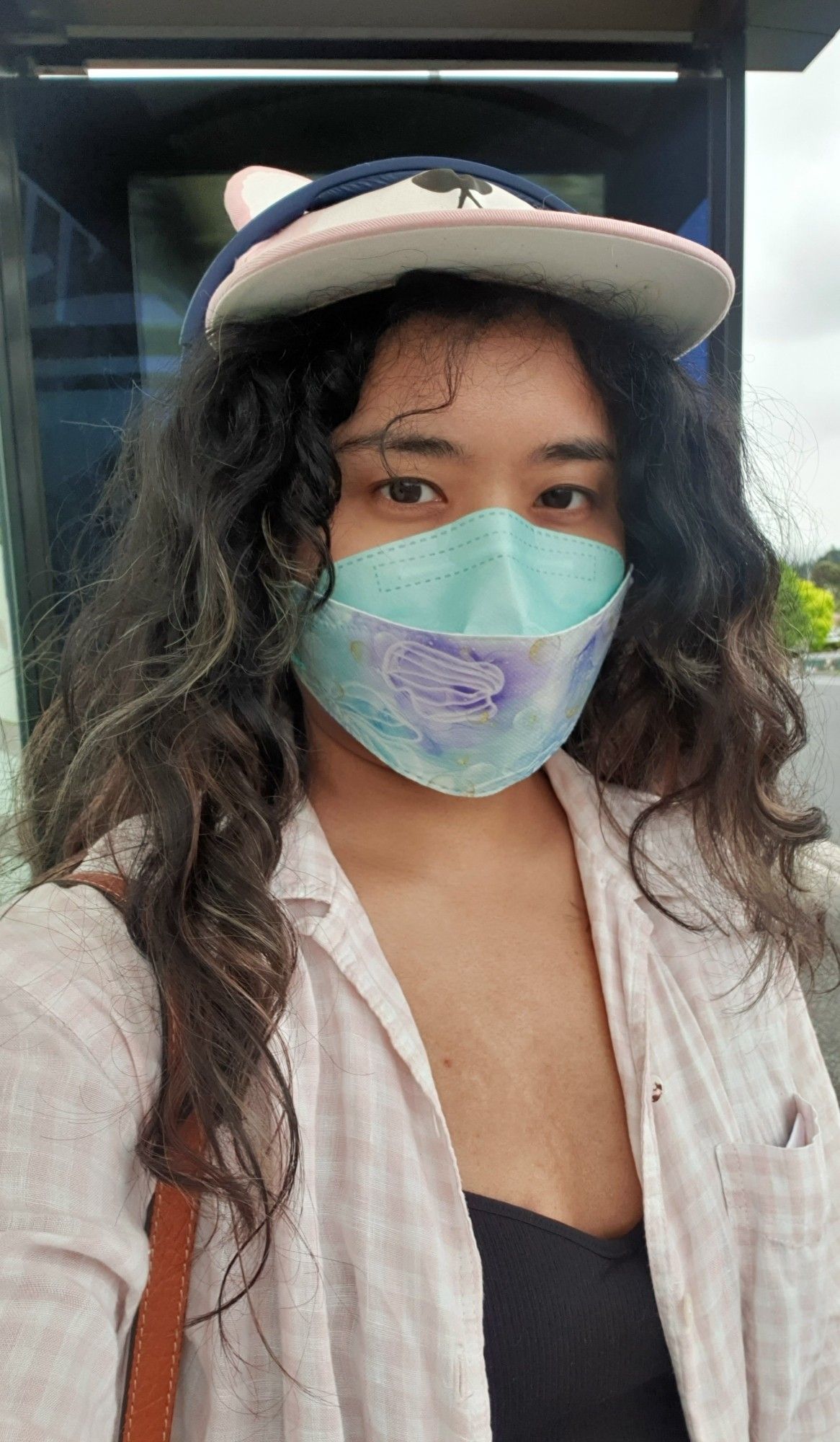 a photograph of me, a brown person with long loose curls. i am wearing a pink snapback with ears, a gingham button down, a black tank top, and a pastel mask with jellyfish printed on it.