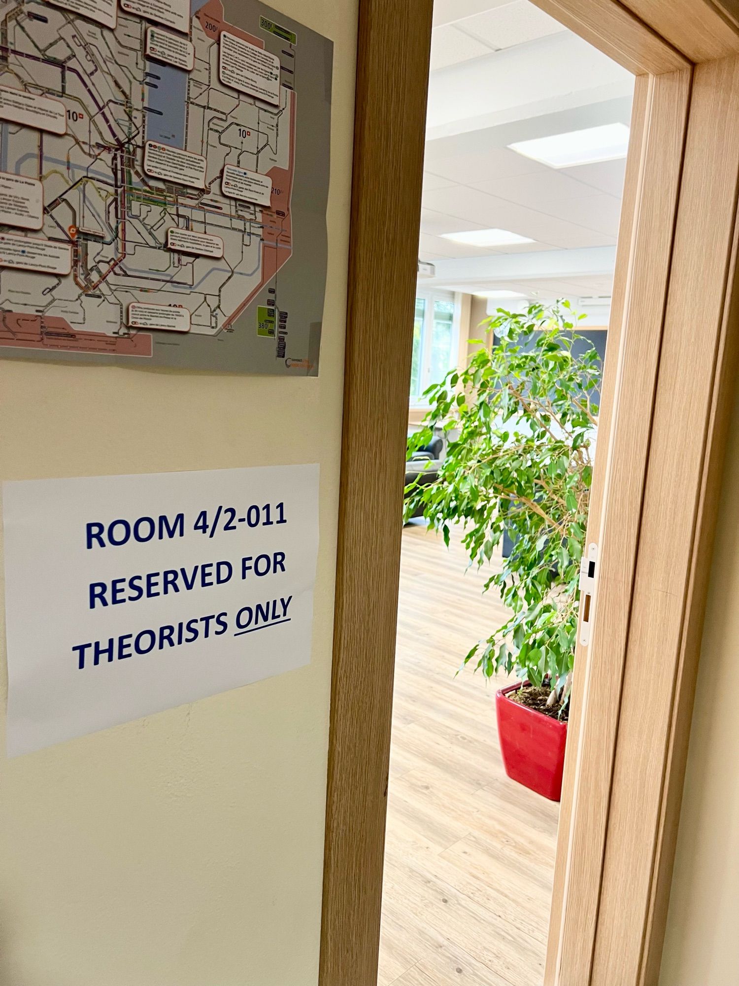 A view into a brightly lit lounge room that contains a ficus plant and some blackboards along the far wall. Next to the doorway under a metro map there is a sign saying “ROOM 4/2-011 RESERVED FOR THEORISTS ONLY” with the last word underlined