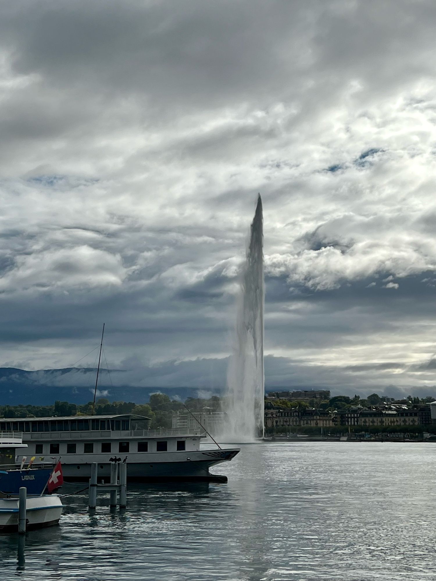 The giant water spout in Lake Geneva climbing high into the sky with extremely dramatic clouds in the background. There are some boats in the foreground and nice hotel-like buildings on the distant shore