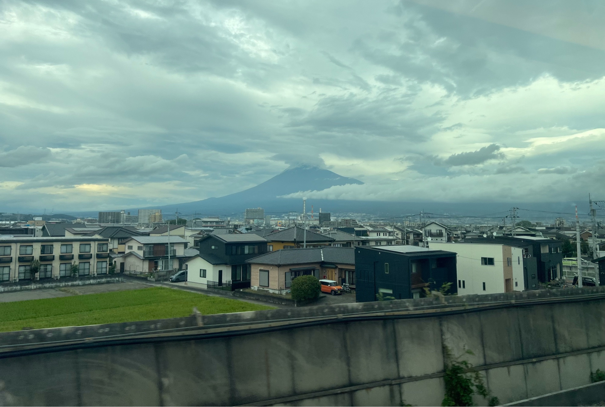 A view of houses and fields and Mount Fuji in the distance partially obscured by large sweeping clouds