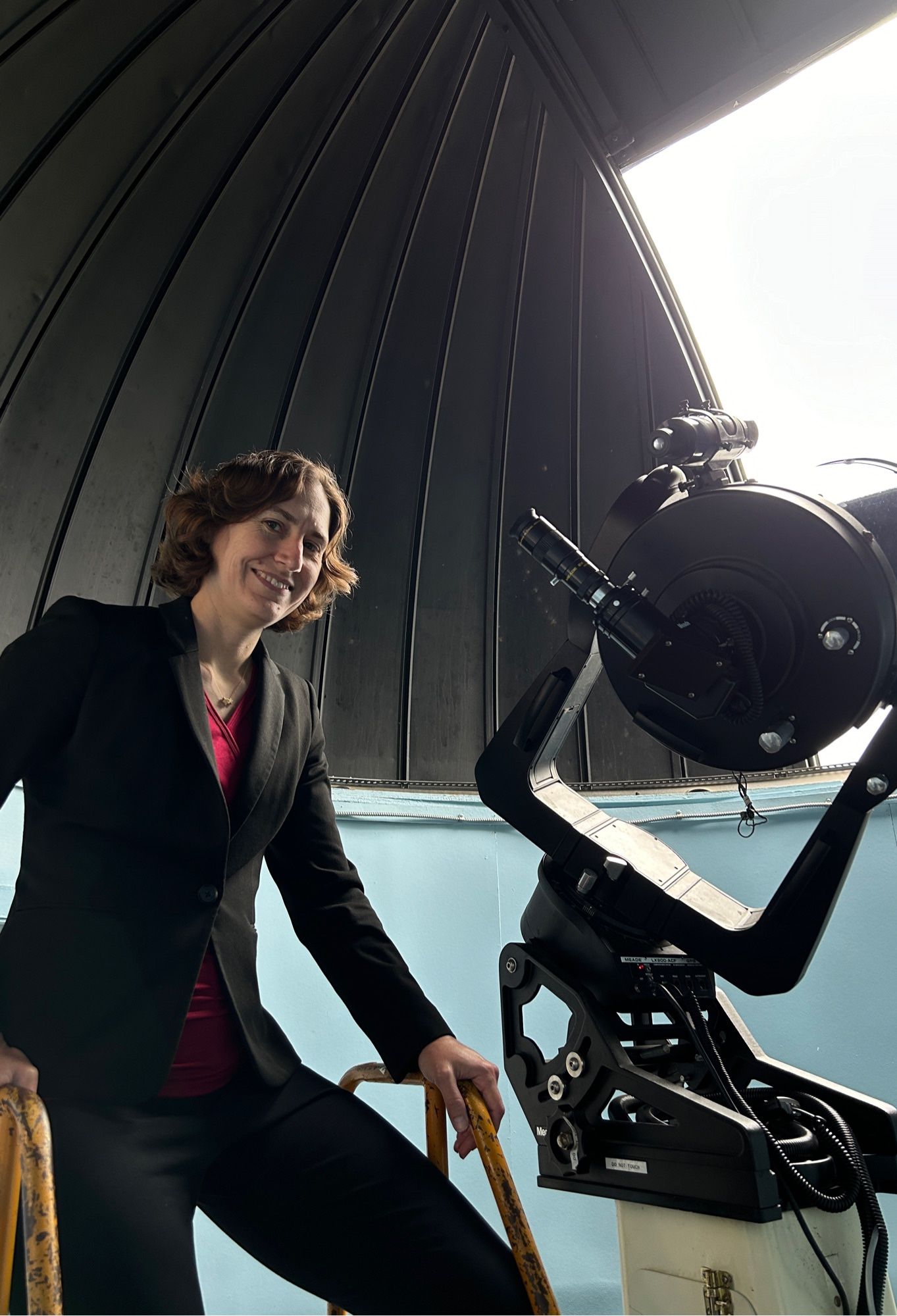 A photo of me standing on a short ladder next to a telescope in a telescope dome. It’s daytime so you can tell it’s all for show but the telescope is legit used for observing