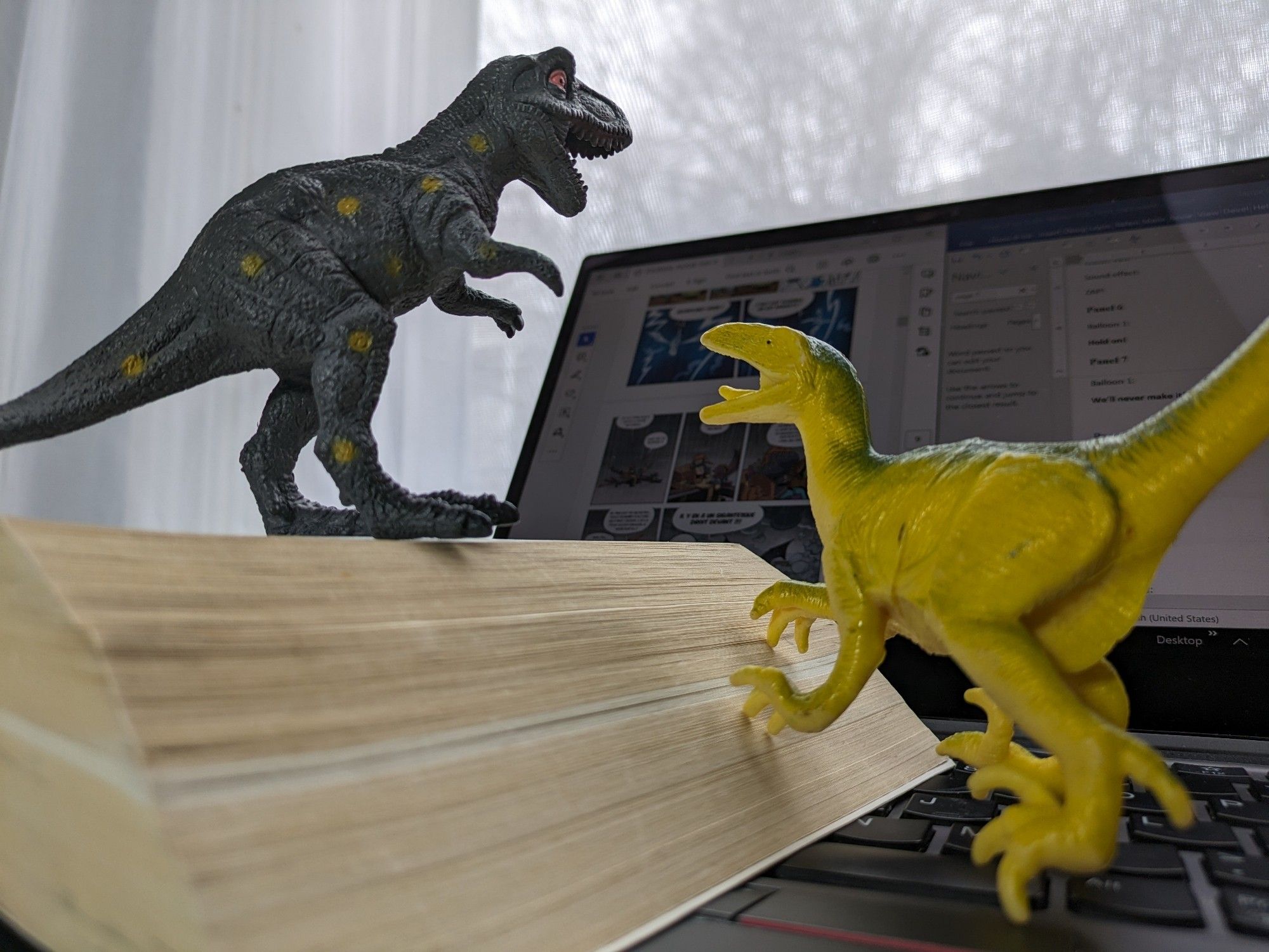 Two toy dinosaurs on top of a dictionary in front of a laptop screen. There's a blurred graphic novel and a translation script on the screen.