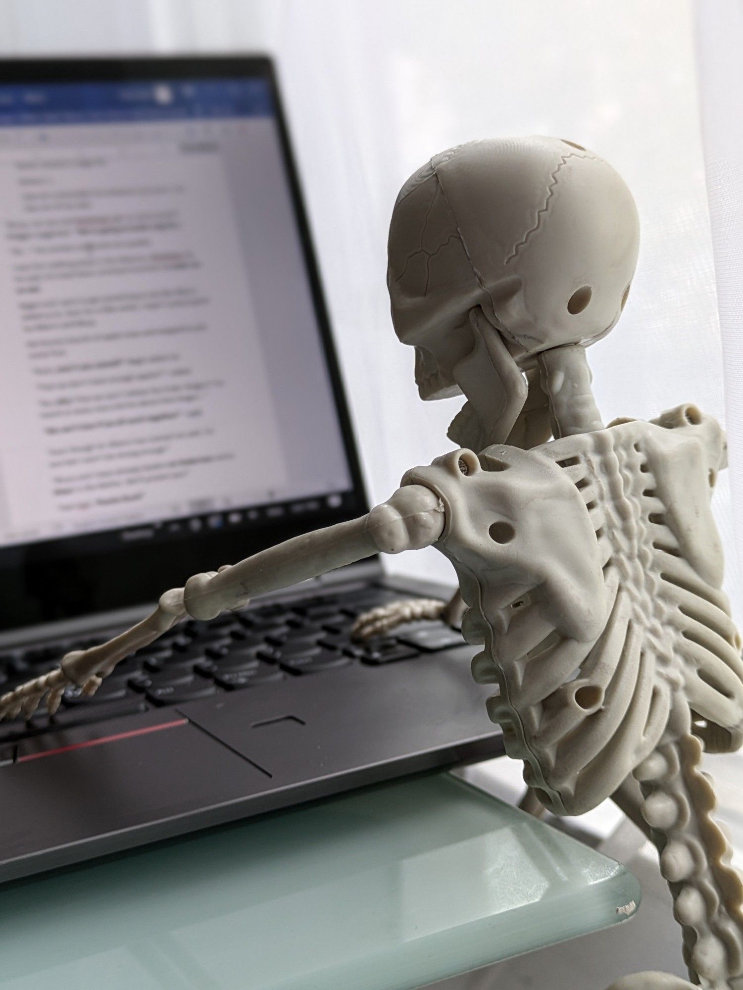 A small toy skeleton in front of a laptop with a blurred Word doc on the screen