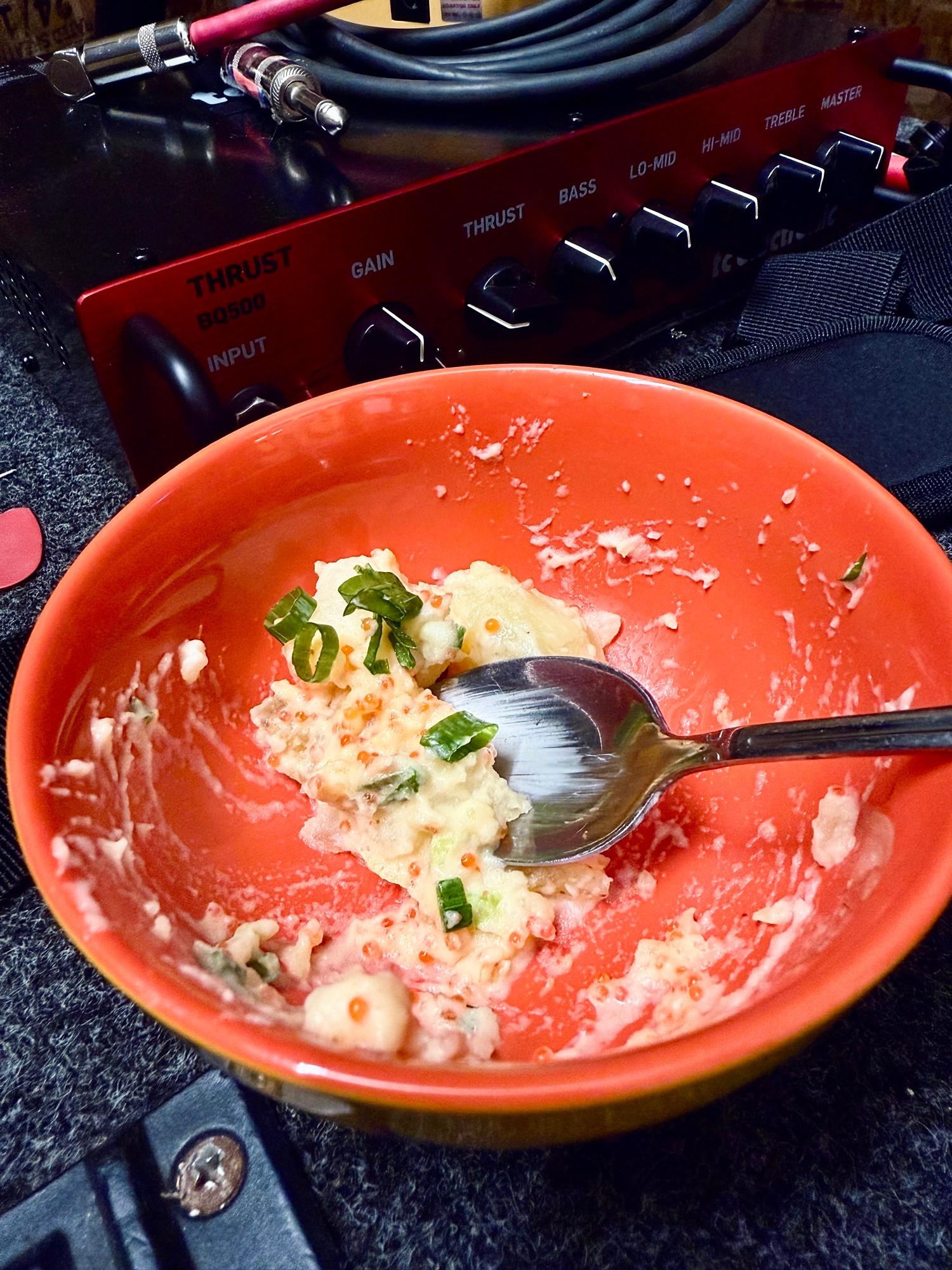 A little orange bowl with spoon and a few bites of mentaiko potato salad left in it, sitting on top of a bass cabinet in front of an amp