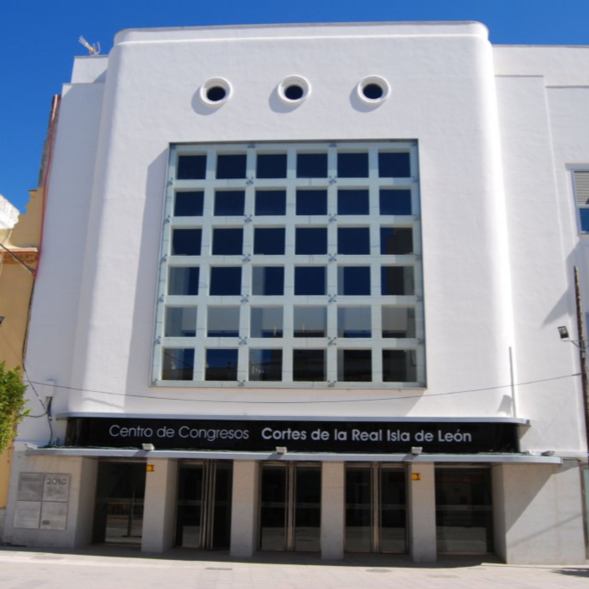 Fachada del Centro de Congresos y Exposiciones «Cortes de la Real Isla de León». Es un edificio blanco, de 3 plantas, con un diseño moderno, aunque restaurado de una edificación clásica. Fachada blanca, 3 grandes puertas de acceso, una cuadrícula de ventanas en el centro y tres ojos de buey o claraboyas en la parte superior. Más bonito que un sanluis de palo.