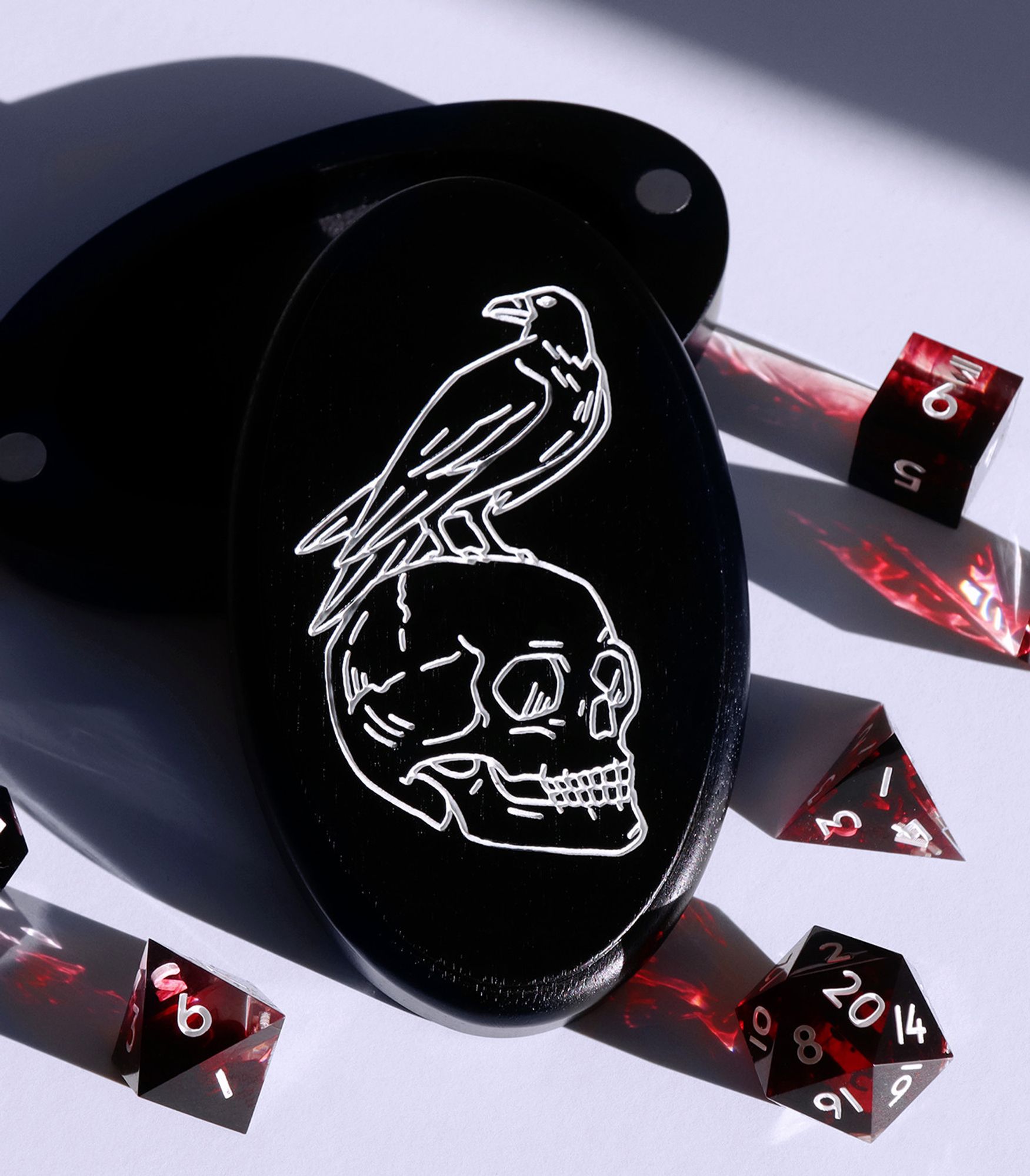 Memento Mori dice vault on a white background surrounded by bloody red dice. The vault is a black walnut wood with an etched silver design of a skull with a raven sitting on top.