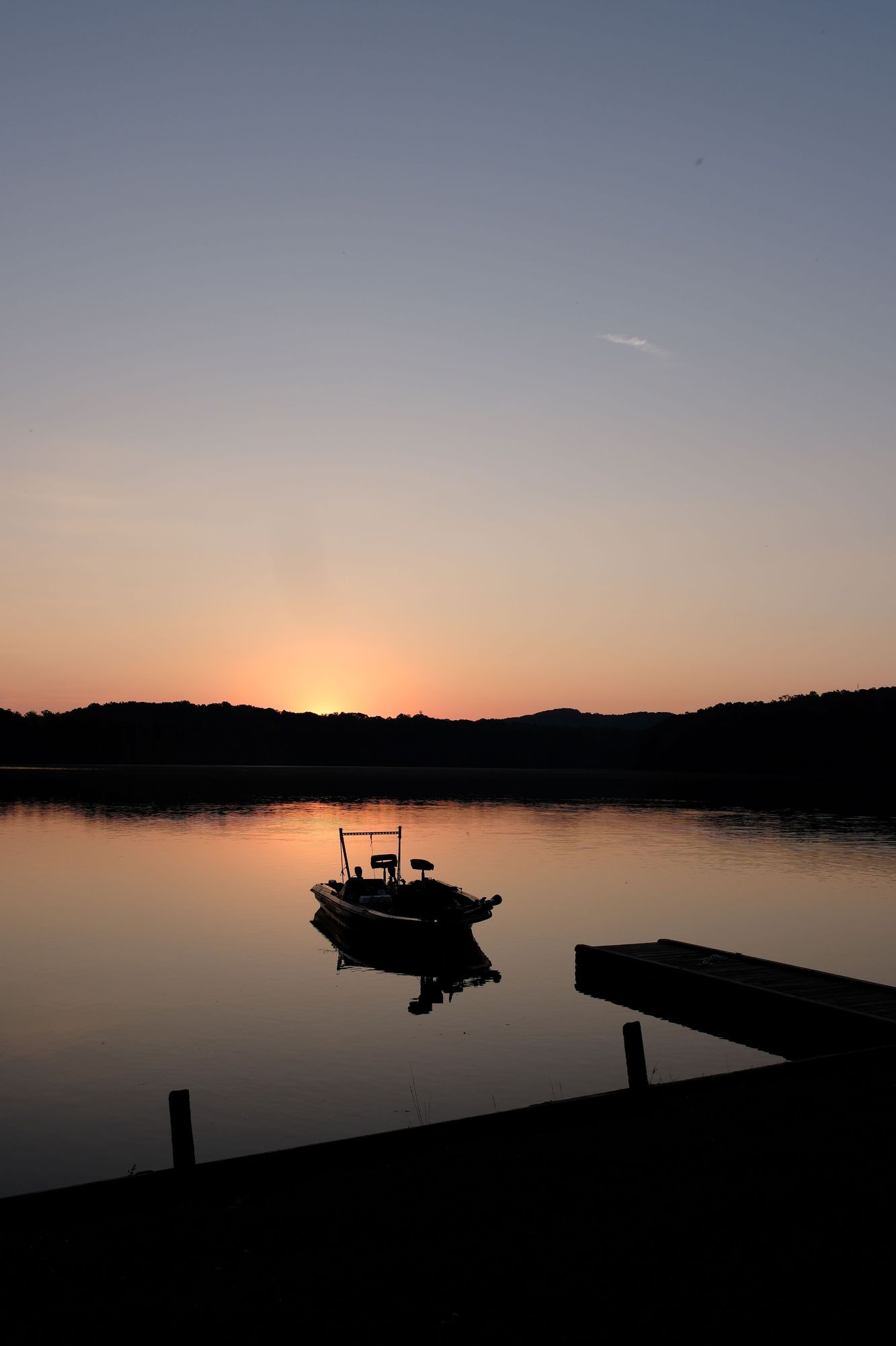 The golden hour as the sun is peeking above the woods on the shore of a lake. A boat is floating on the lake, with a wooden jetty on the bottom right.