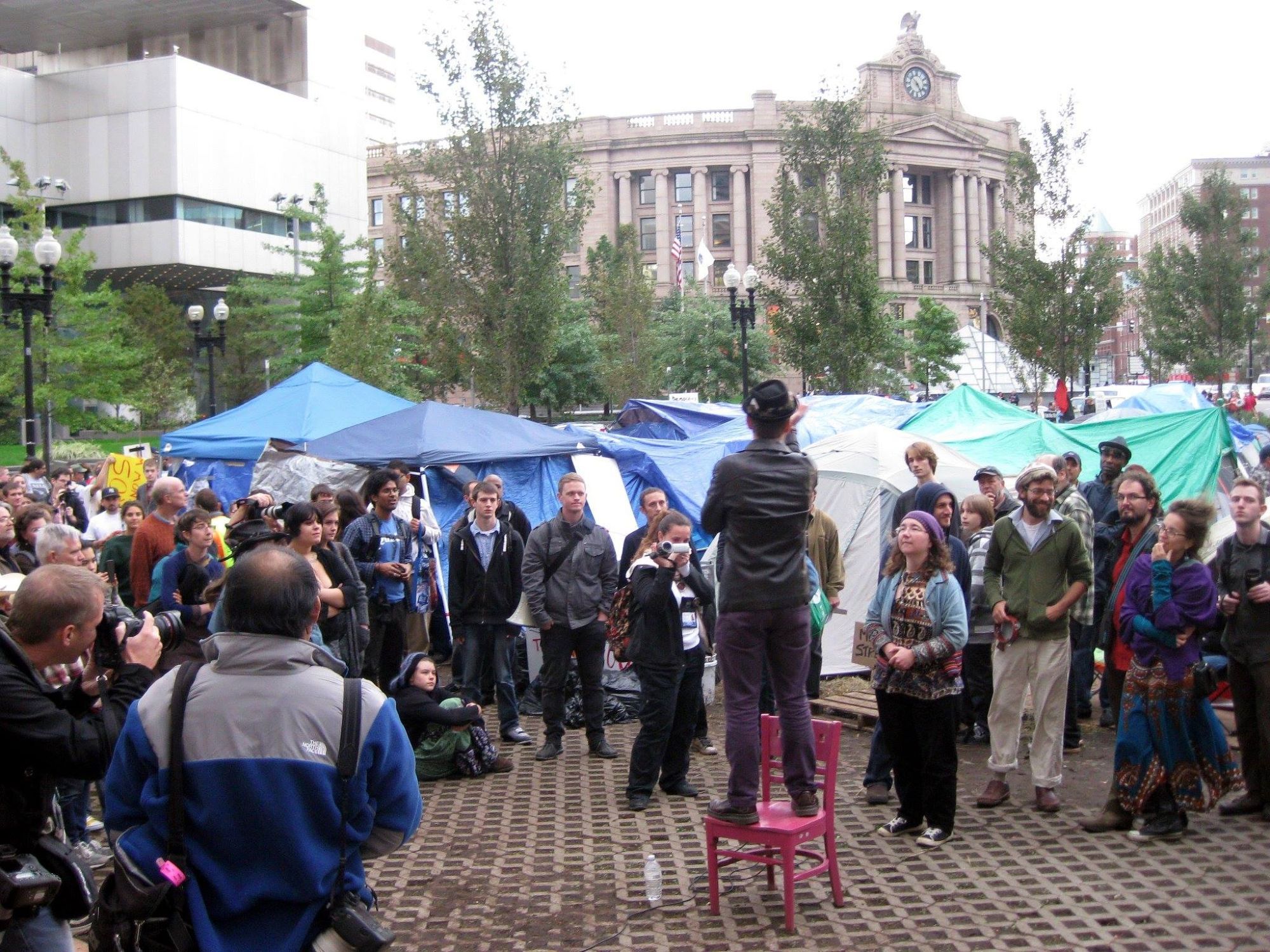 a photo of Dewey Square (the site of Occupy Boston). A speaker stands on a chair, surrounded by a crowd. Beyond them are tents.