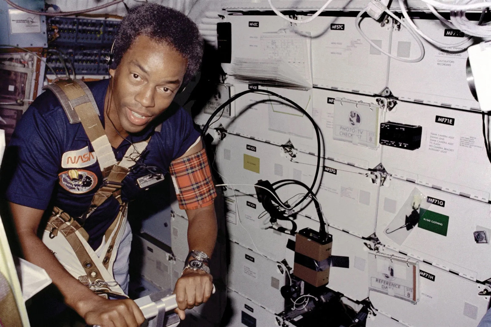 A Black man floating in zero gravity on a spaceship. He’s looking at the camera and he’s wearing a NASA shirt.