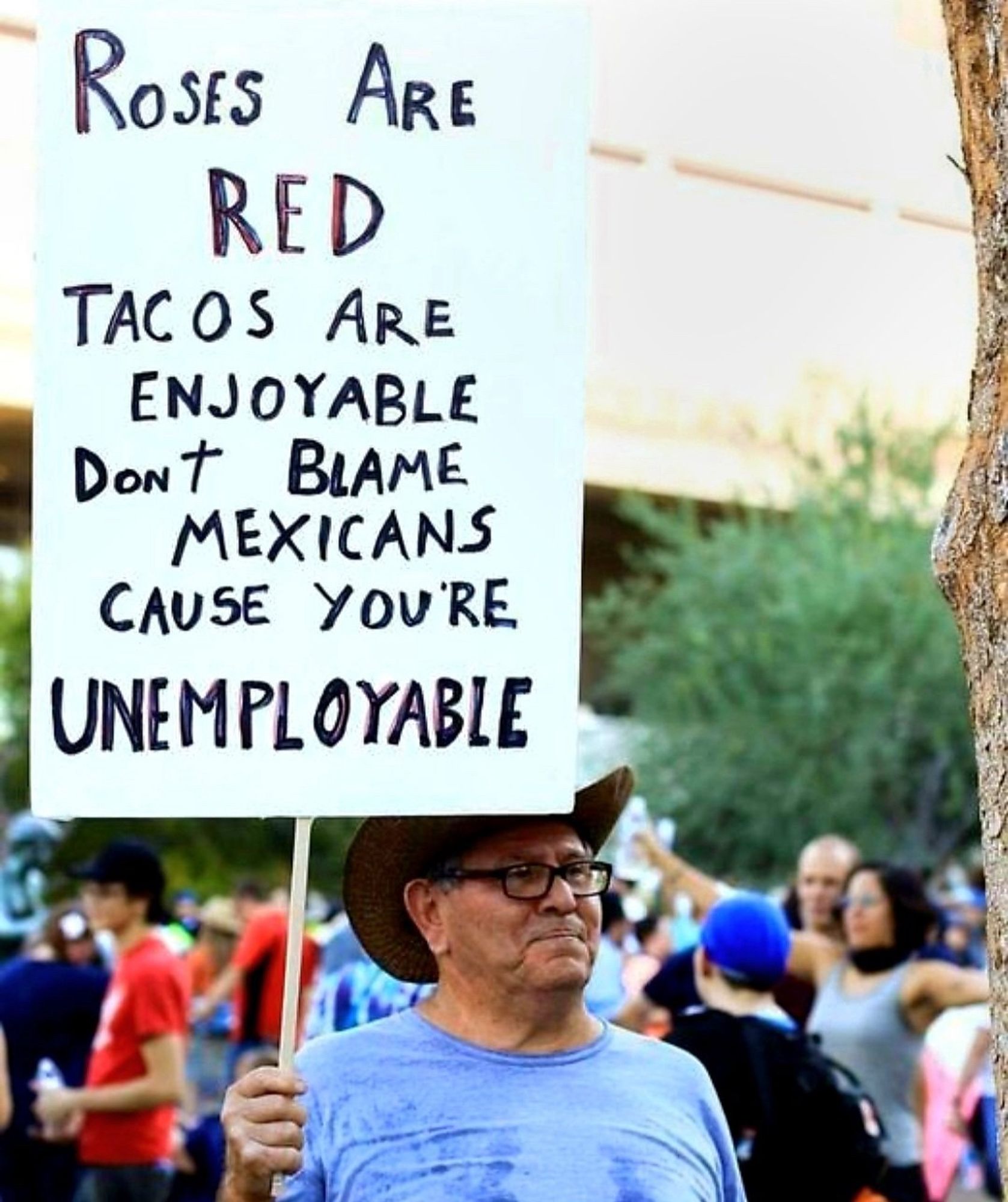 A man holding a protest sign that says:

ROSES ARE RED
TACOS ARE ENJOYABLE
DONT BLAME MEXICANS
CAUSE YOU'RE UNEMPLOYABLE