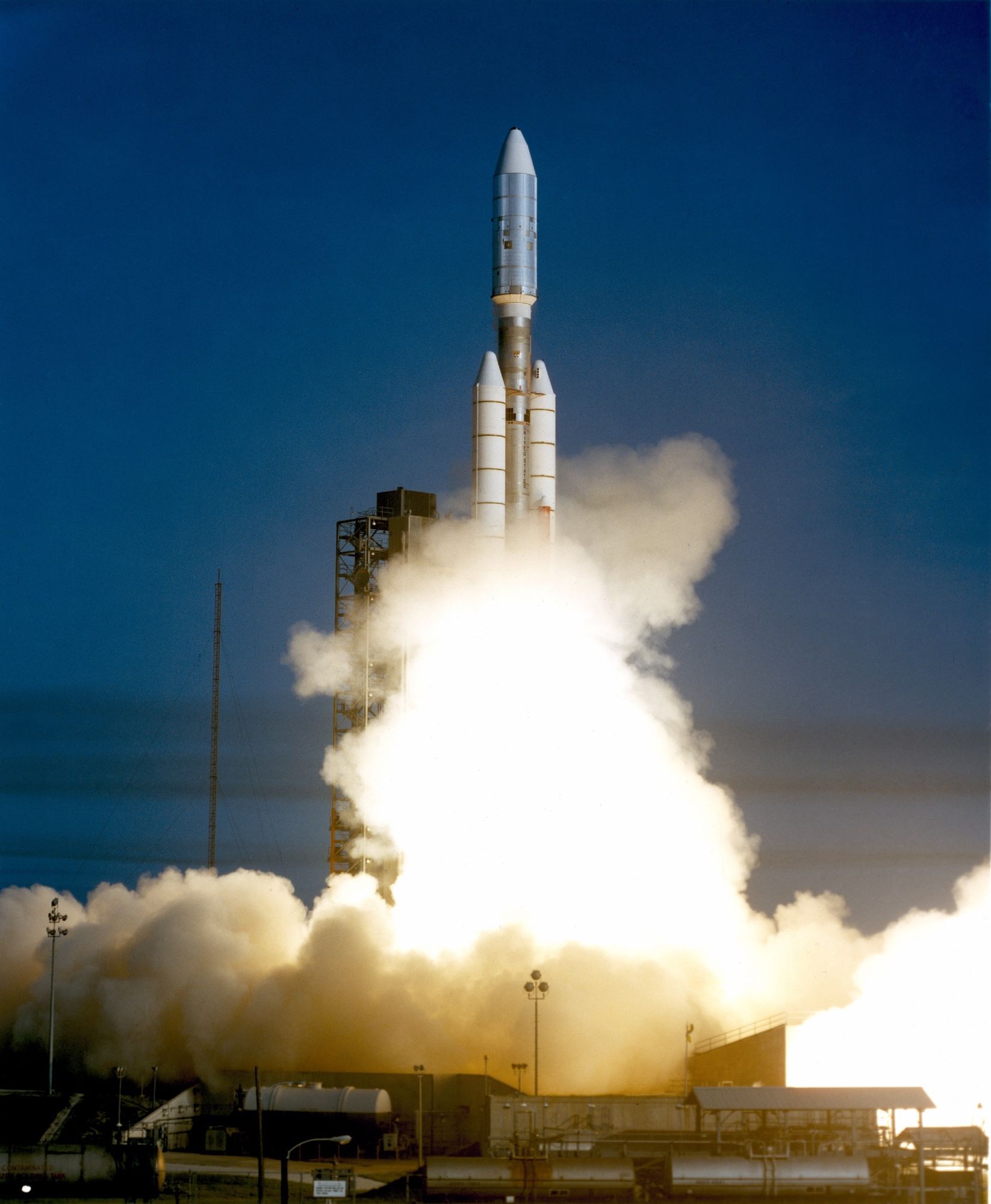 Color photo of the Voyager 1 launch. The probe sits atop a rocket mounted on a larger rocket with twin solid fuel boosters – a Titan IIIE launch vehicle. The photo is taken just after ignition. The bottom of the rocket has lifted up about half the height of the gantry, and is engulfed in bright white flame and smoke.