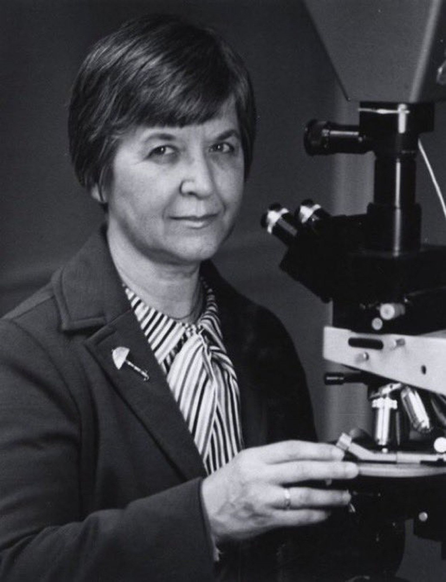 Black and white photo of Stephanie Kwolek standing in front of a microscope. She wears a dark jacket and striped blouse, with a lapel pin that looks like an umbrella. Her dark hair is worn in a short bob and she is looking at the camera.