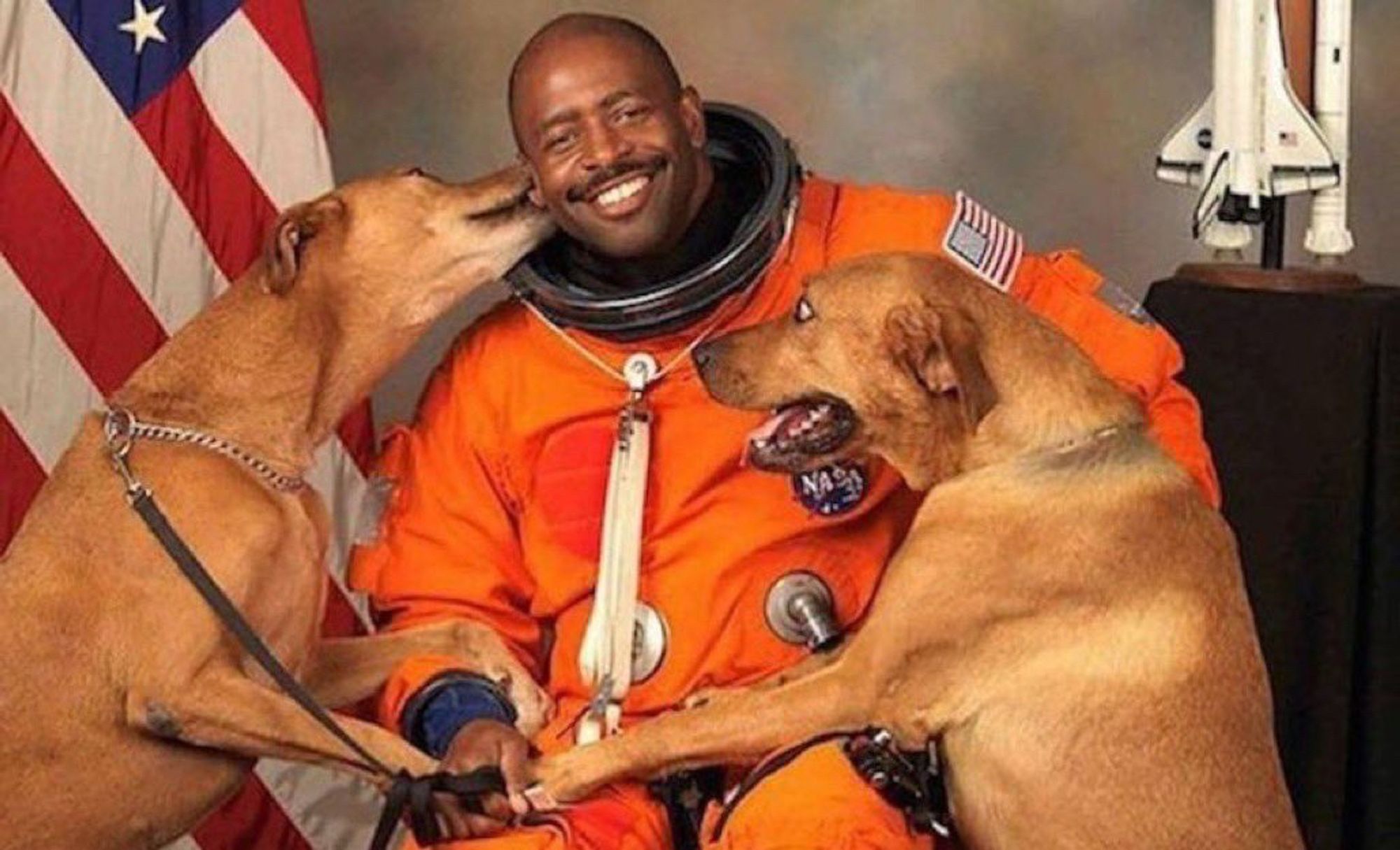 Astronaut Leland Melvin in his orange NASA astronaut jumpsuit, with his two dogs climbing all over him.